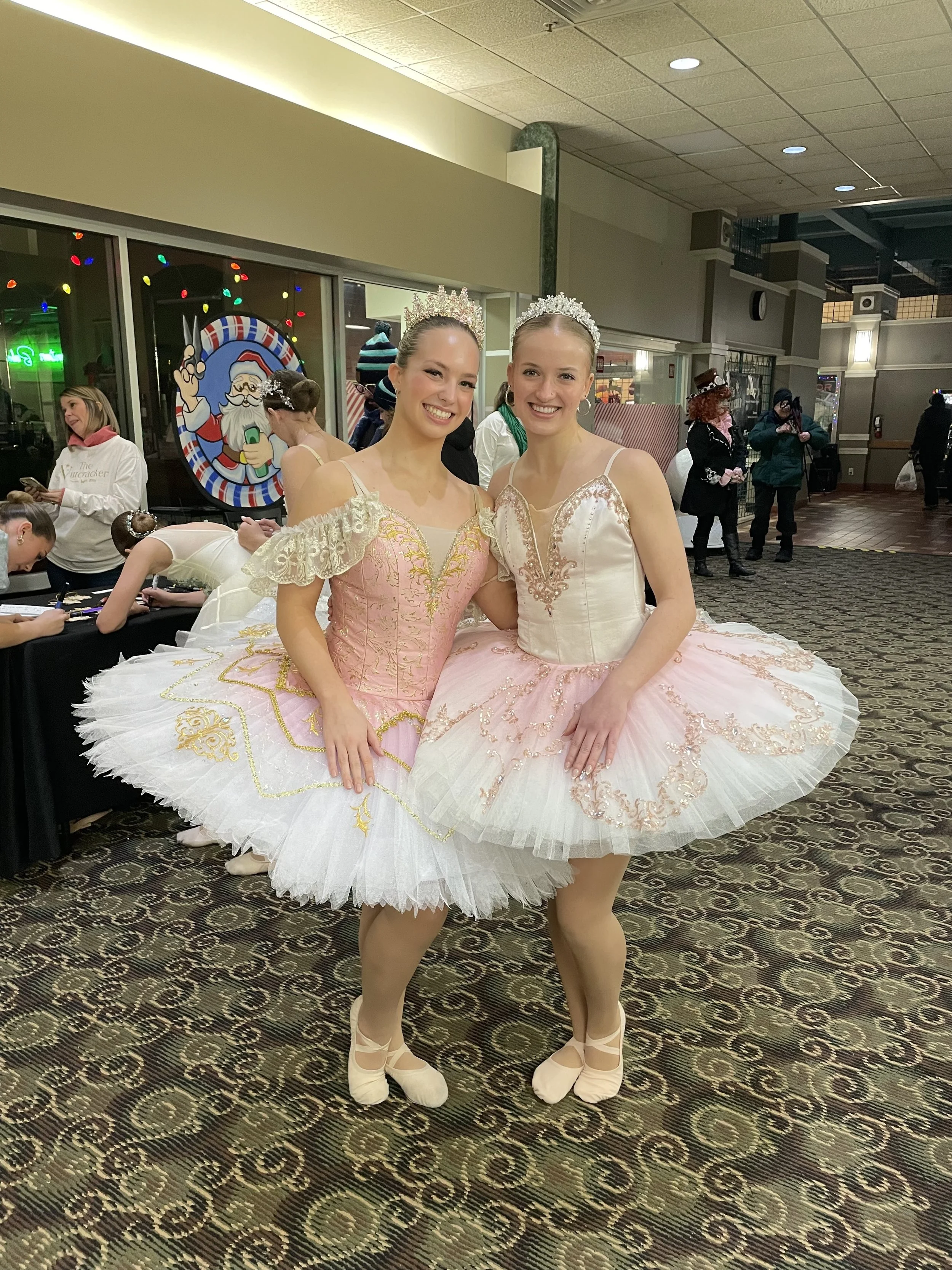 Two ballet dancers in pink and white tutus with tiaras pose together in costume at an indoor event.