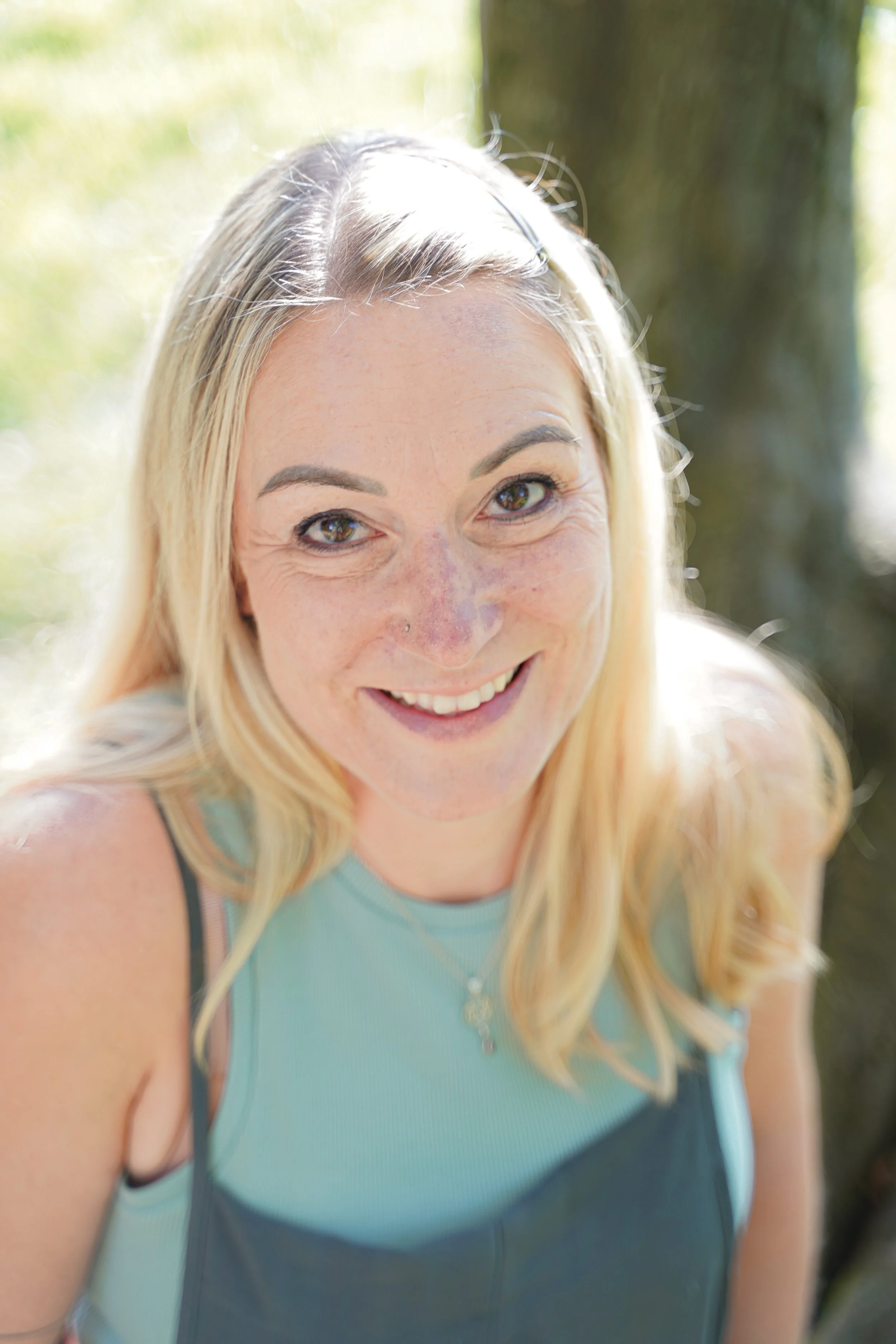 Lächelnde Frau mit blonden Haaren, die draußen im Grünen steht und an der Sonne sitzt.