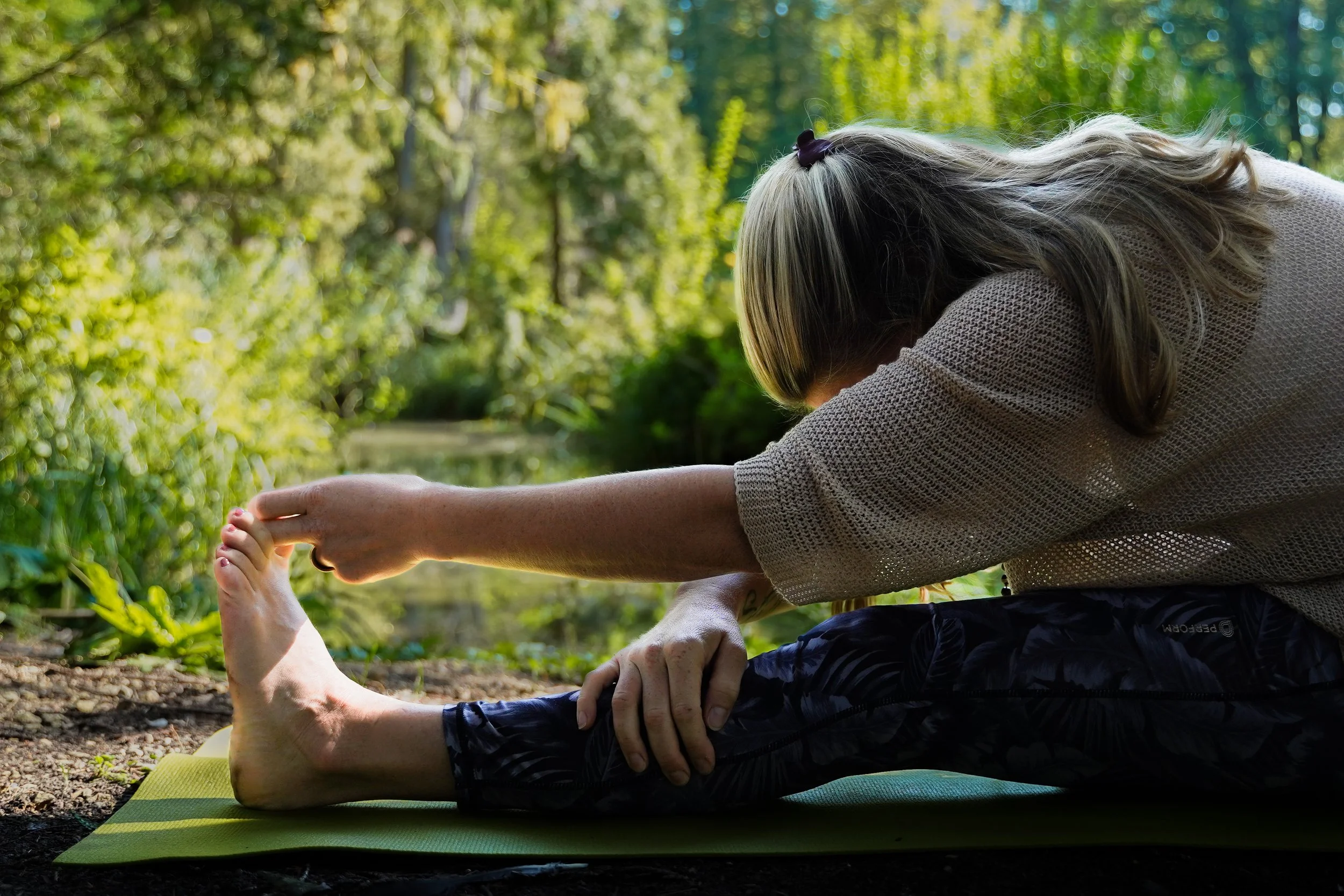 Frau macht Yoga im Freien auf einer grünen Yogamatte in der Natur, umgeben von Bäumen und Wasser.