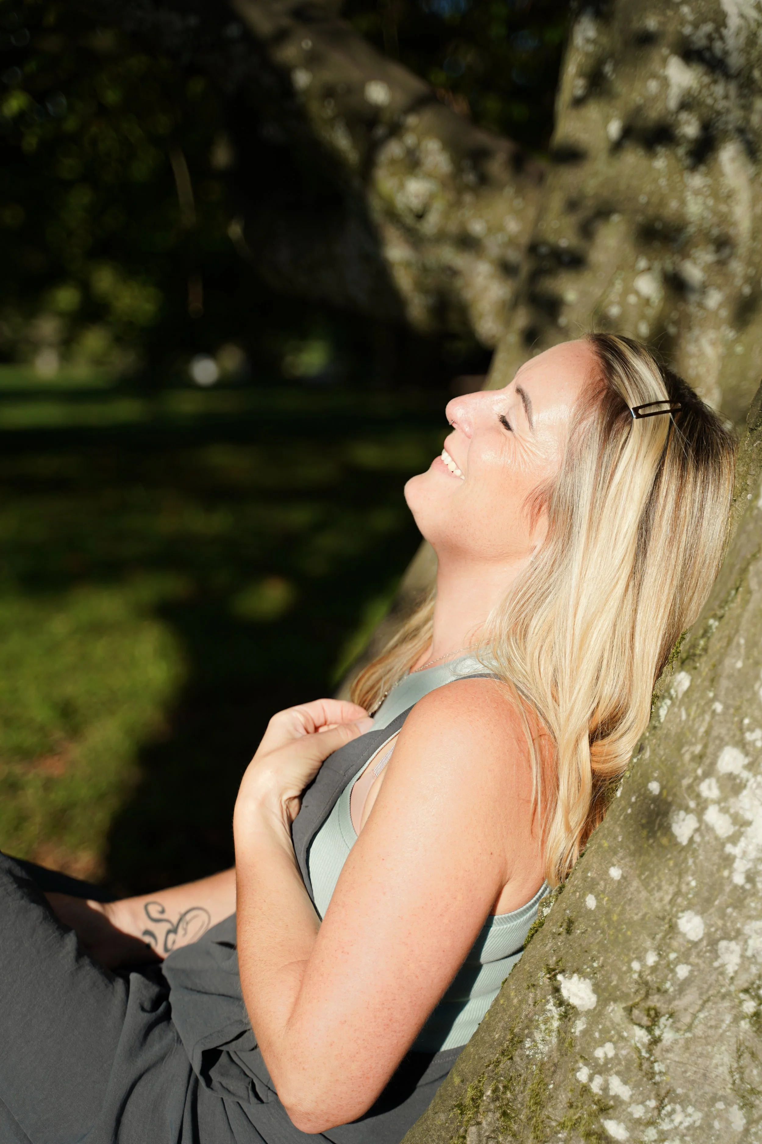Frau sitzt entspannt unter einem Baum, lächelt und genießt die Sonne im Park.