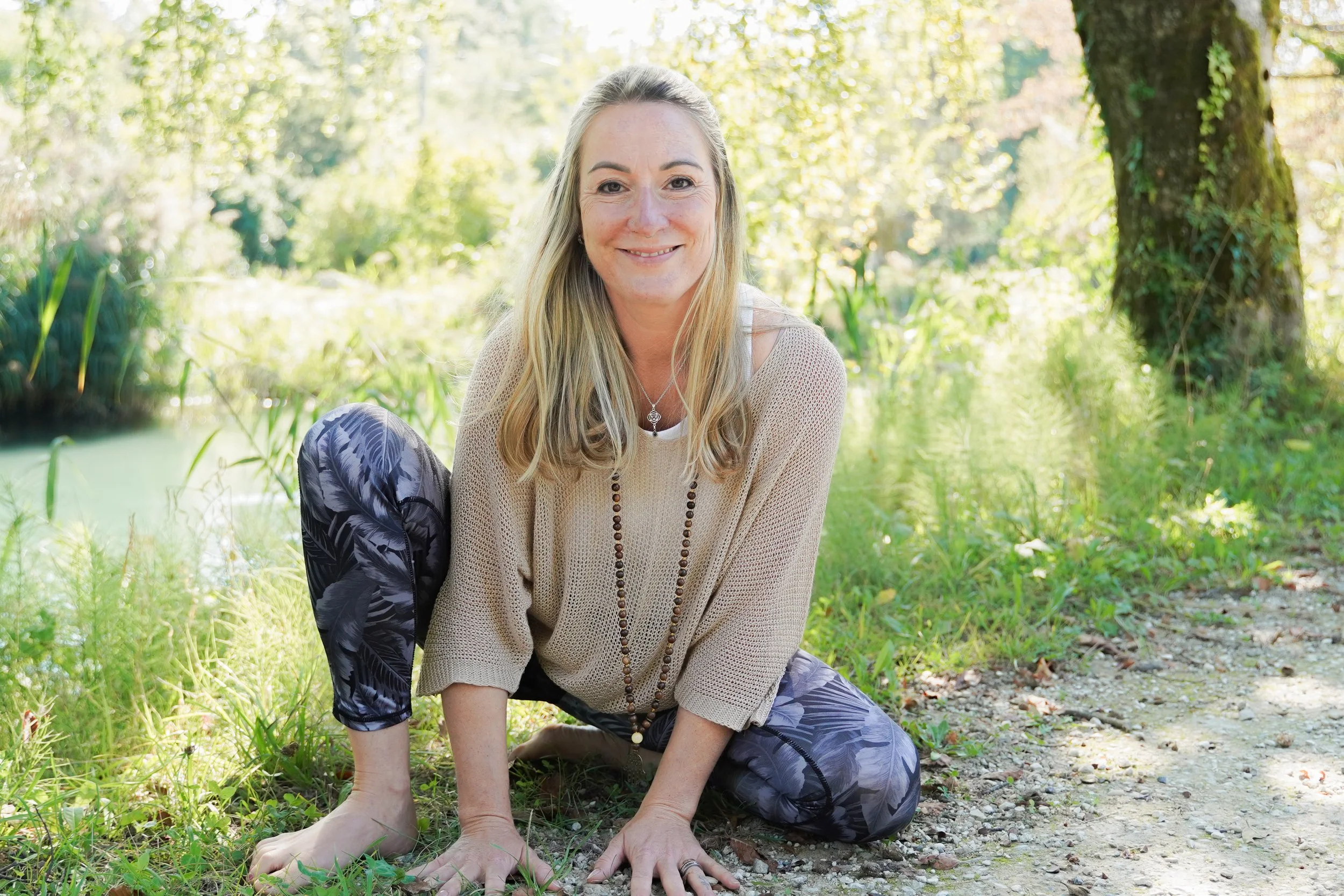 Eine lächelnde Frau mit langen blonden Haaren, die in einer natürlichen, grünen Umgebung auf einem Weg im Knien sitzt.