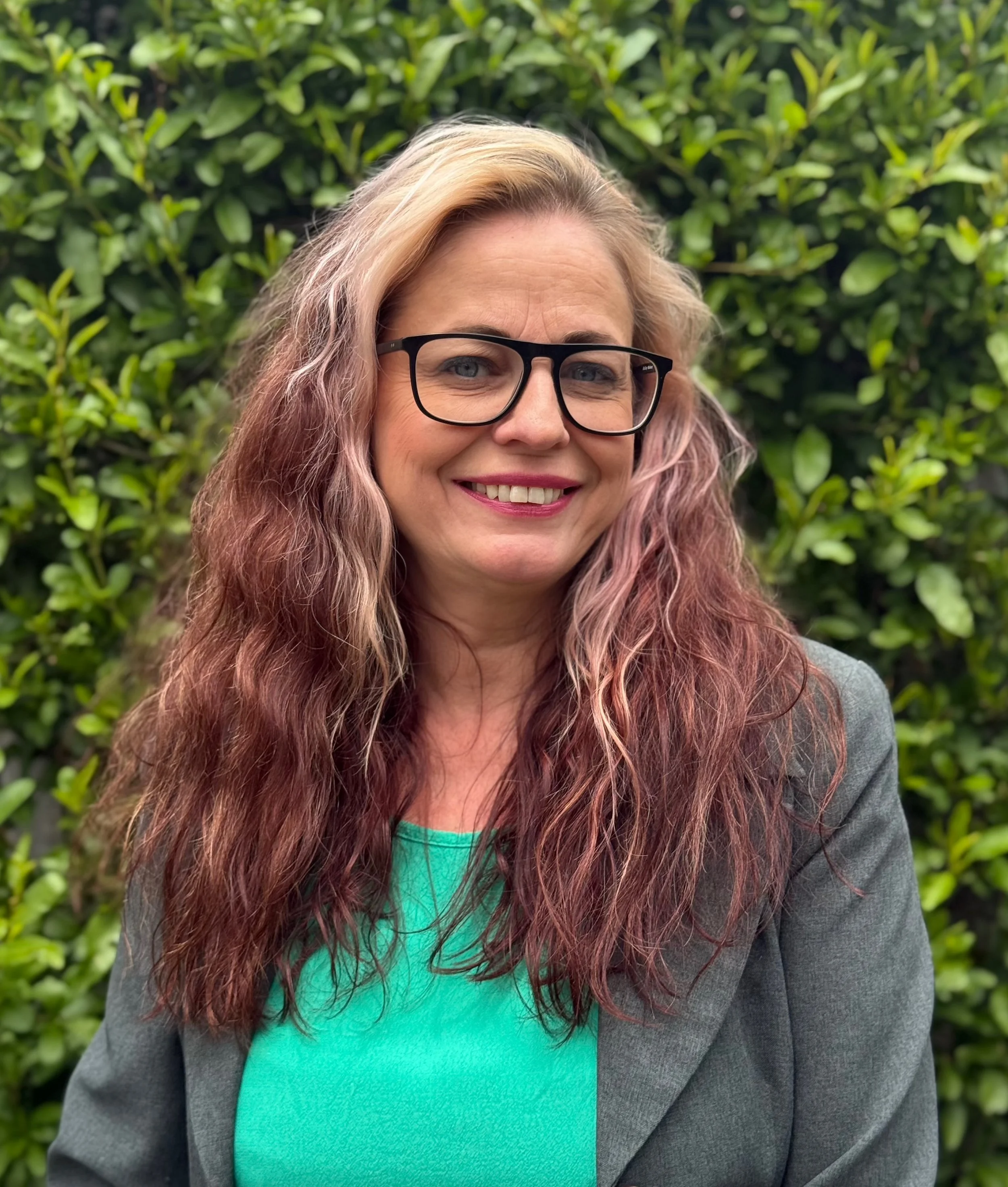 A woman with wavy, shoulder-length pink and blonde hair, wearing black glasses, a gray blazer, and a turquoise top, standing outdoors in front of green shrubbery, smiling.