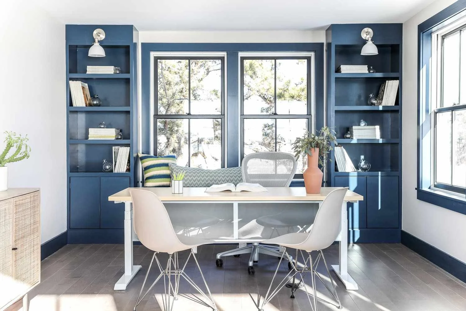 Home office with a desk, four white chairs, a blue wall with built-in shelves, large windows, and plants.