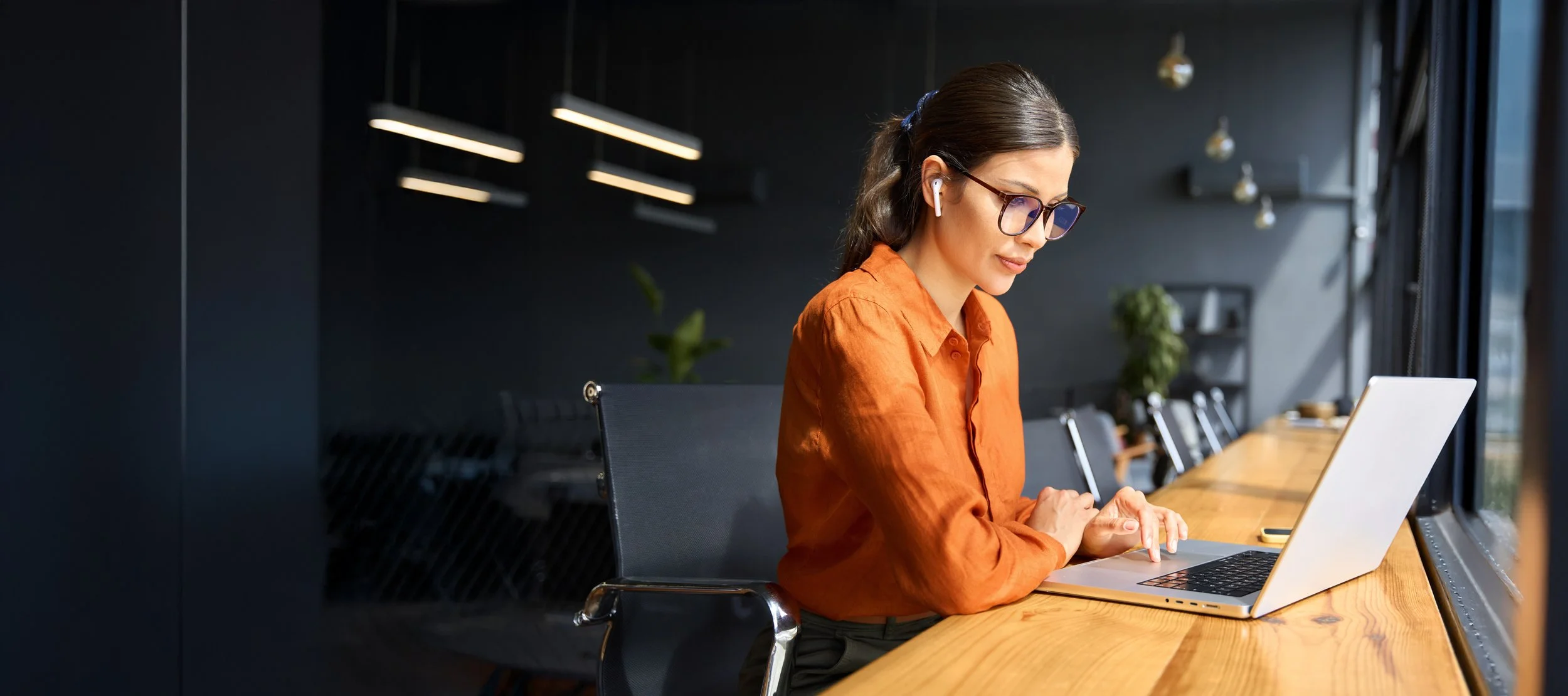 A woman with brown hair, glasses, and wireless earbuds, sitting at a wooden table in a modern, dark-colored room, using a laptop while looking out the window.