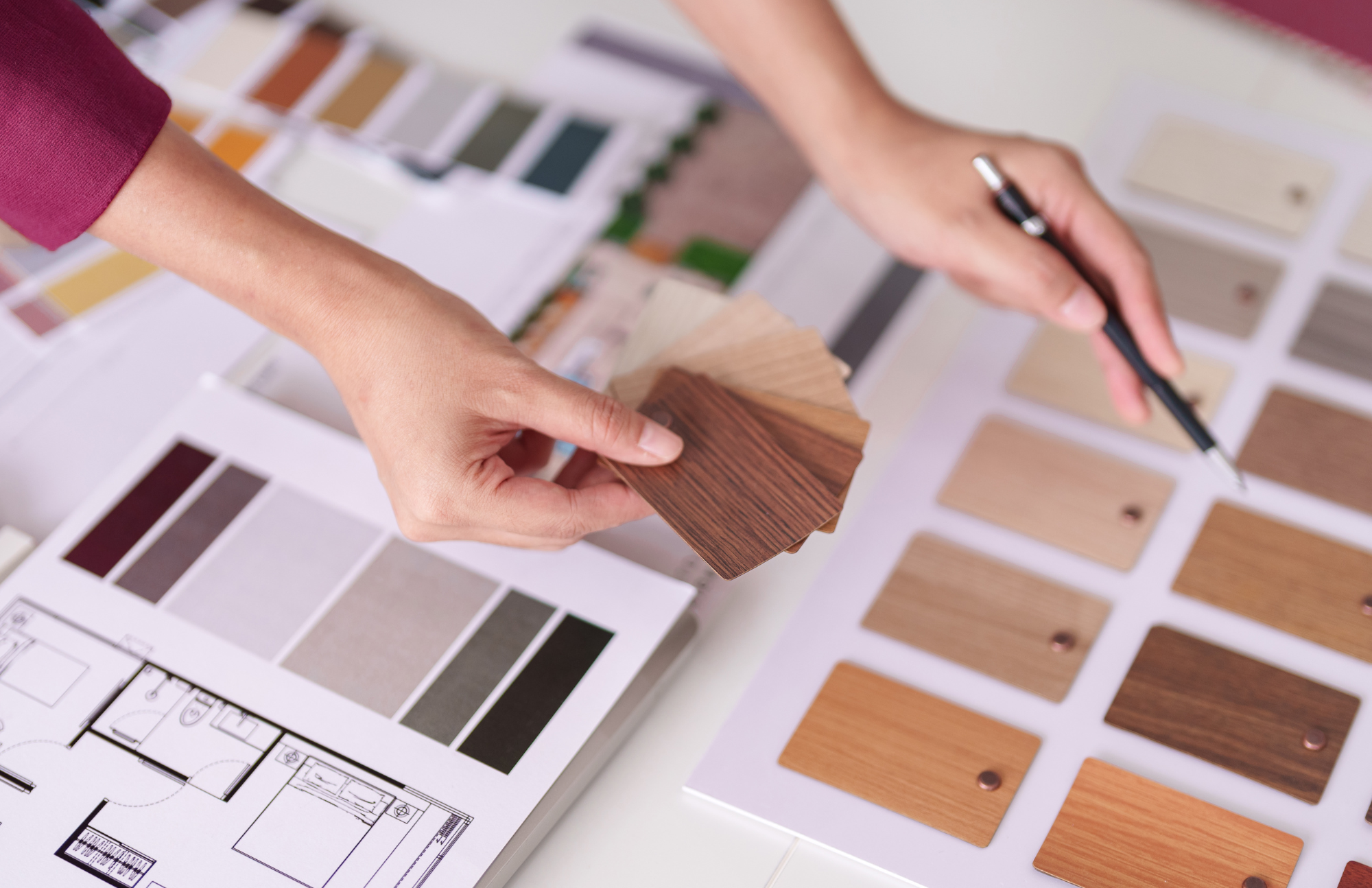 Person holding wood finish sample fan deck and pointing with a pen at a color palette on a table with multiple color sample sheets, design plans, and fabric samples.