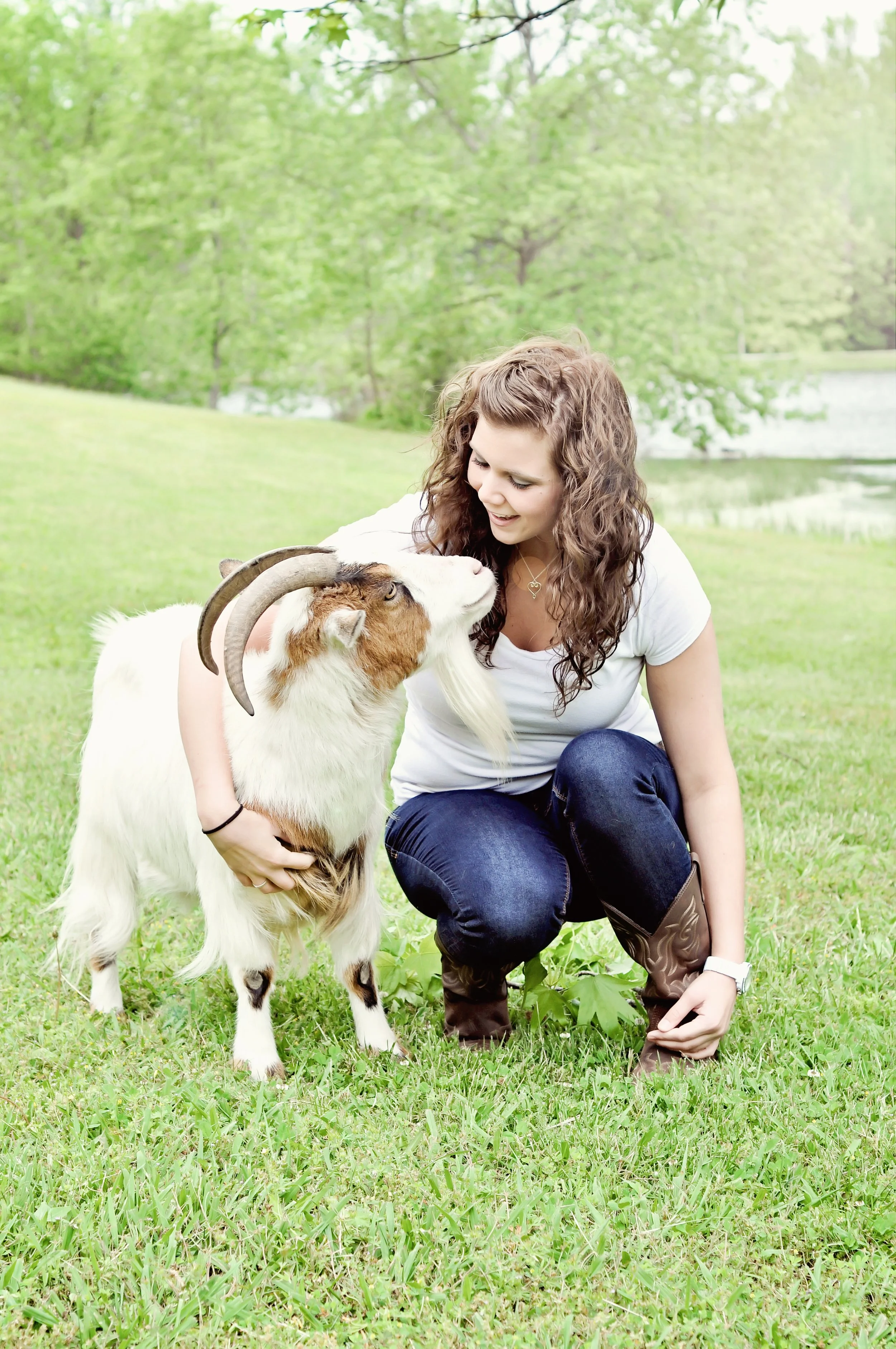 A young woman kneeling on grass and hugging a goat with curved horns in a park near a lake, surrounded by green trees.