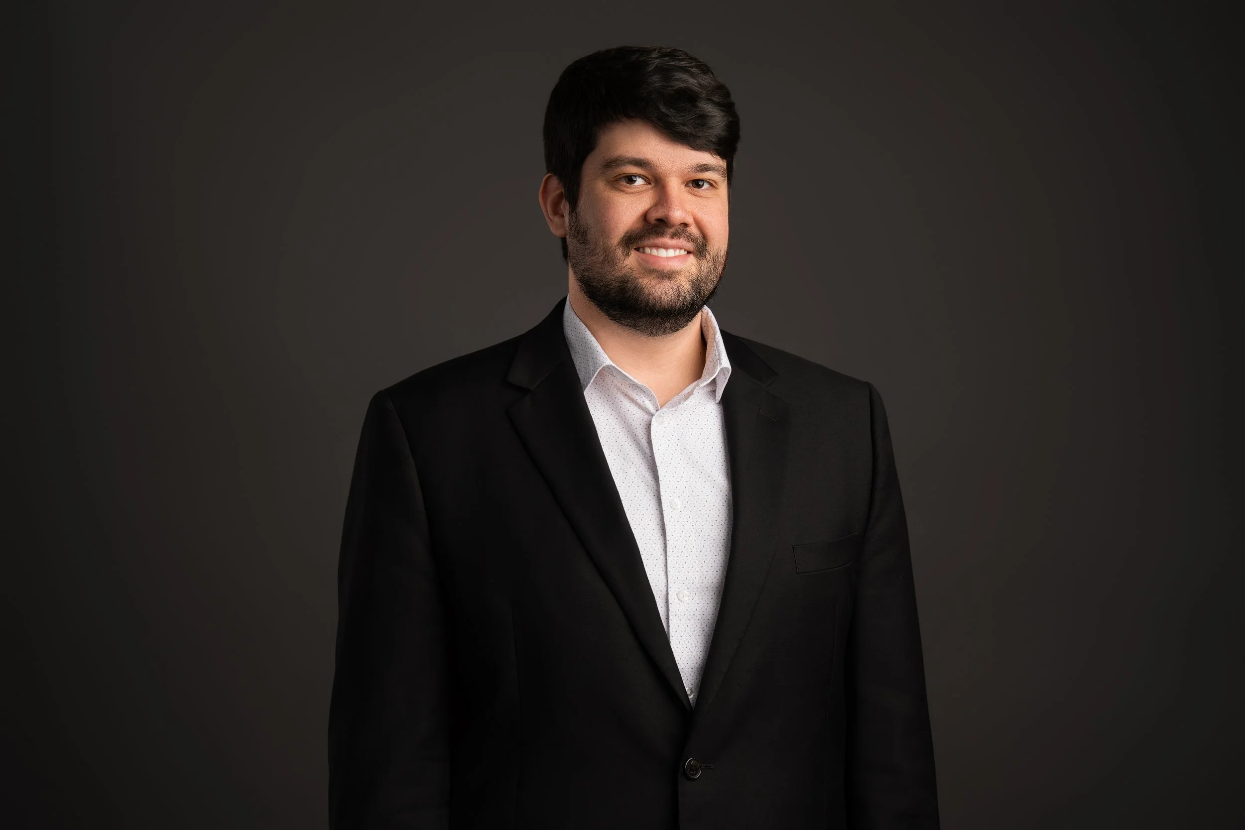 A man with dark hair and beard wearing a black suit jacket and white shirt, smiling against a dark gray background.