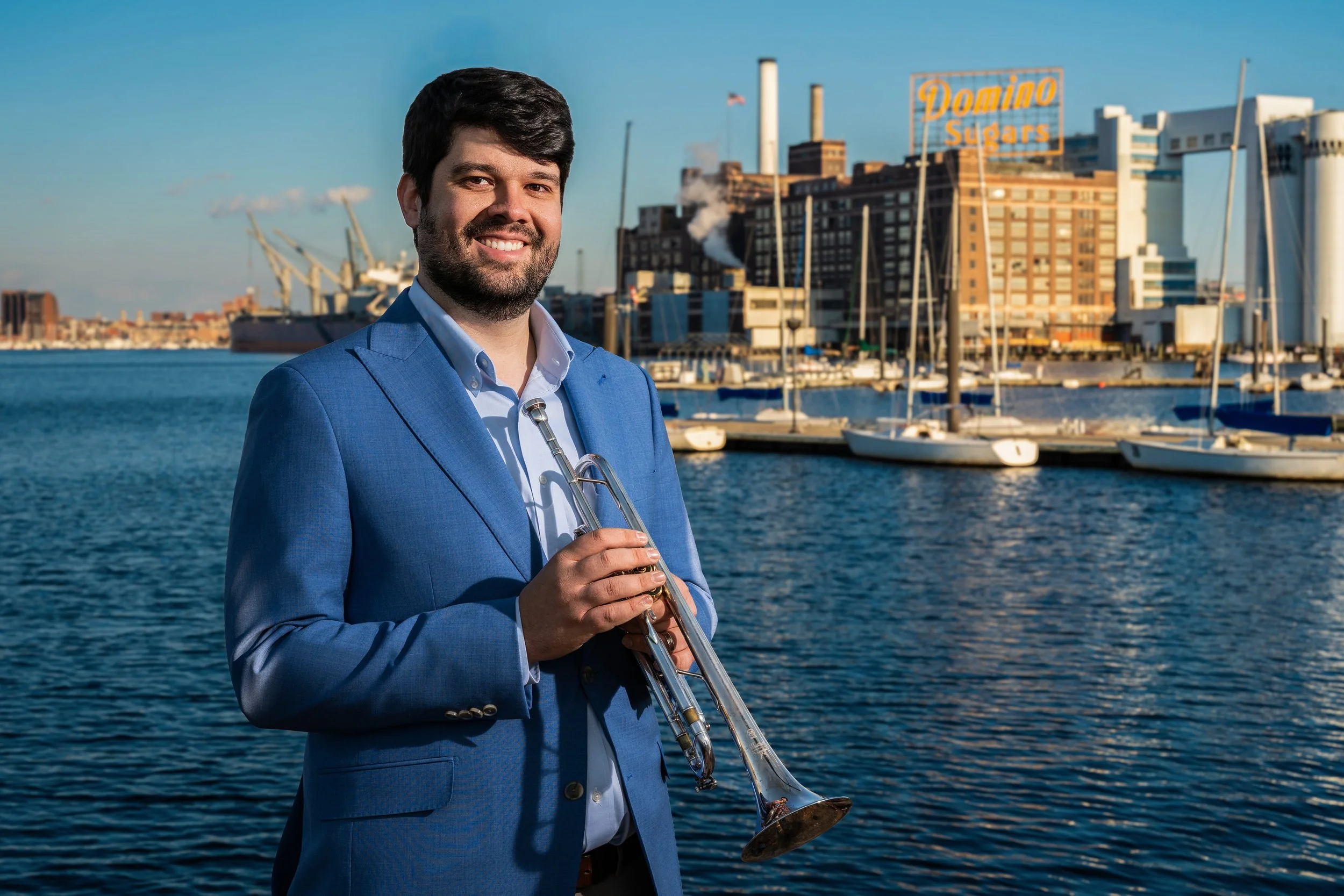 Man in a blue suit holding a trumpet near a marina with sailboats, industrial buildings, and a sign that reads 'Domino Sugars' in the background.