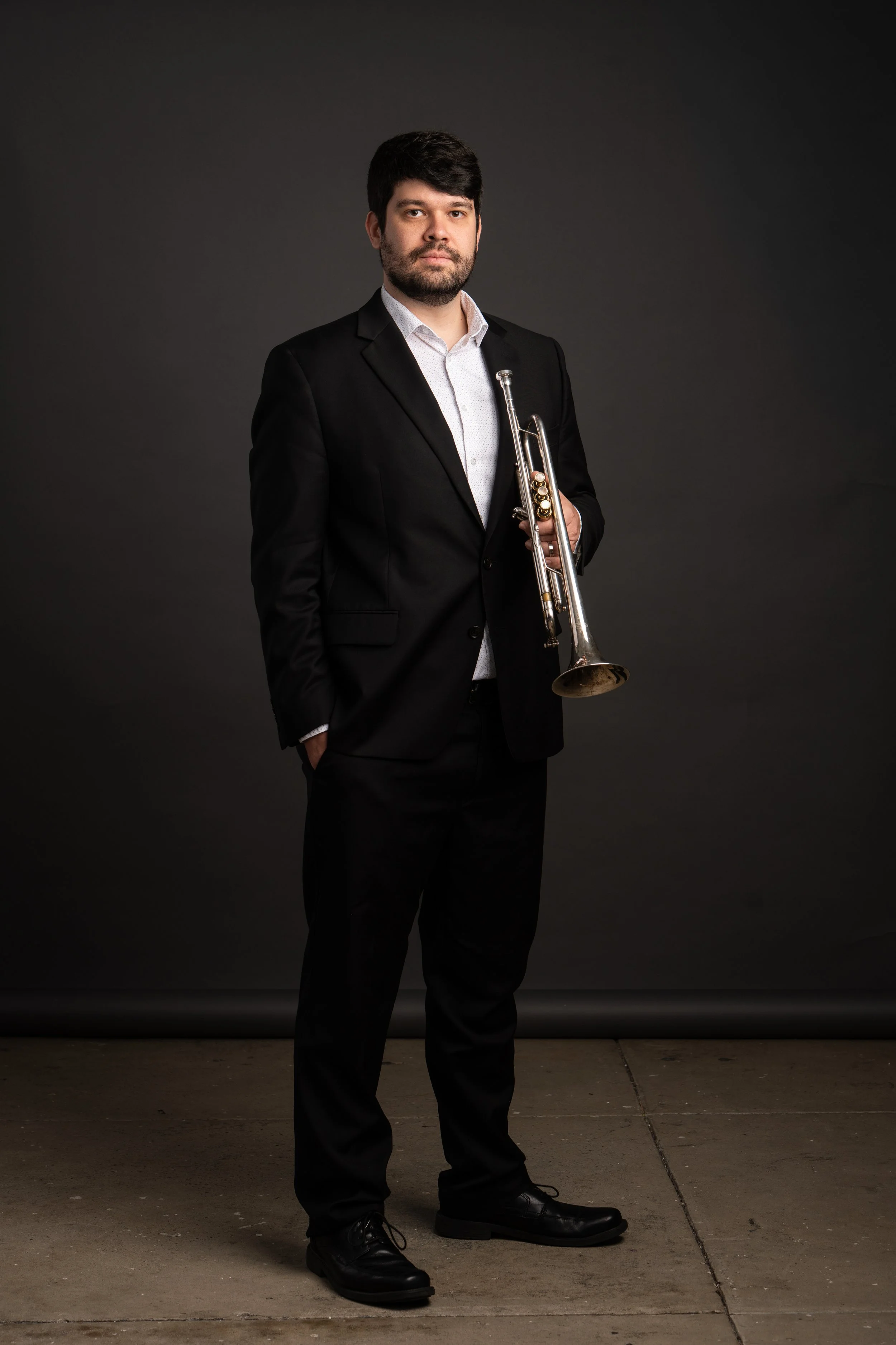 A man in a black suit holding a trumpet, standing against a dark background.