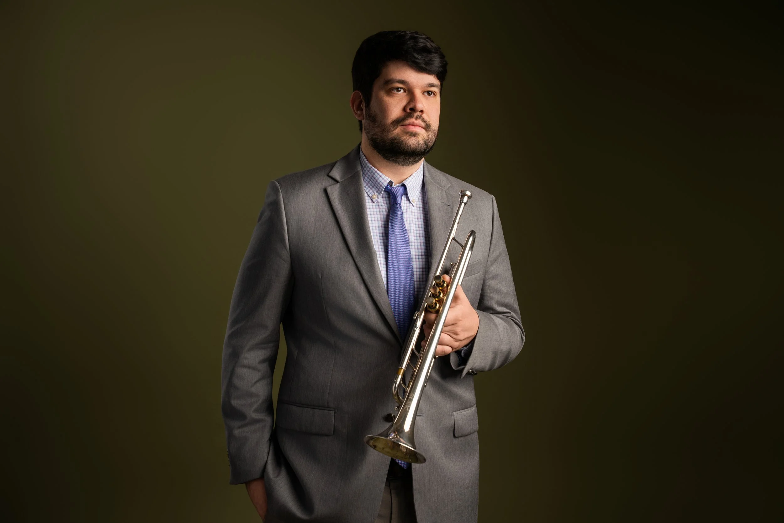 A man in a gray suit holding a trumpet, standing against a dark olive green background.