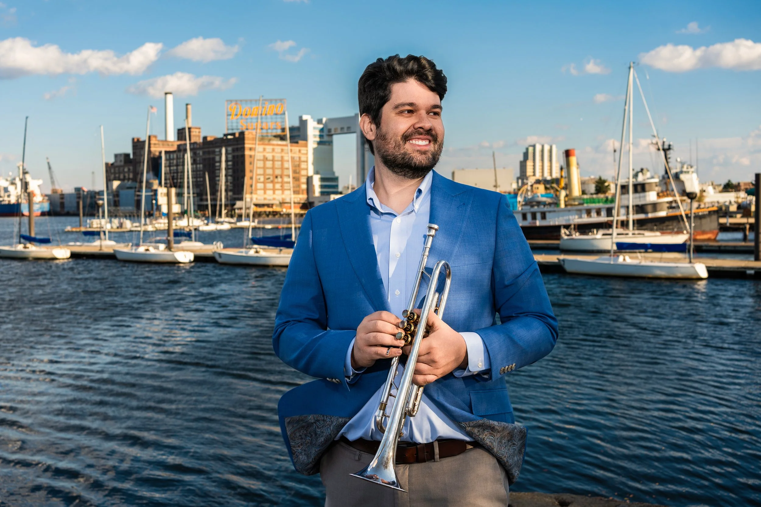 A man in a blue suit holding a trumpet by a marina with boats and city buildings in the background.
