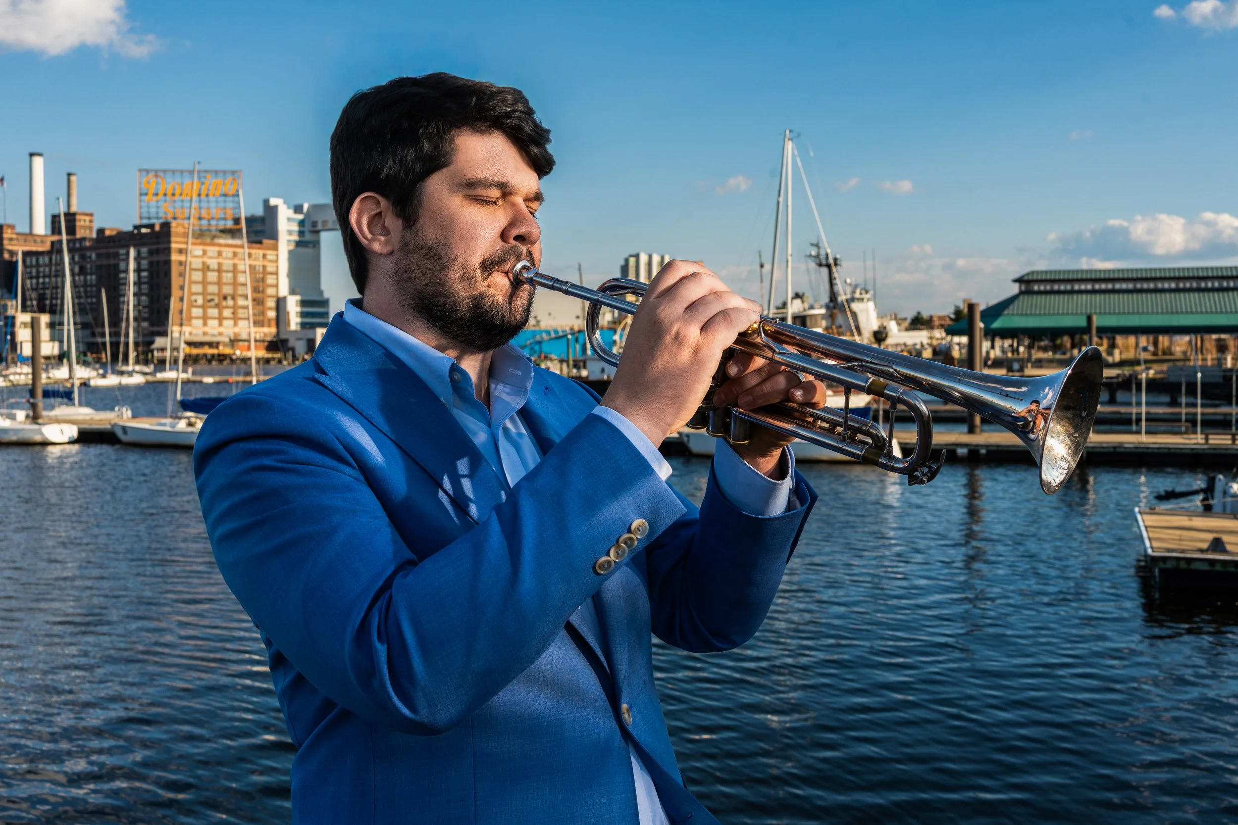 Man in a blue suit playing trumpet near a marina with boats and buildings in the background.