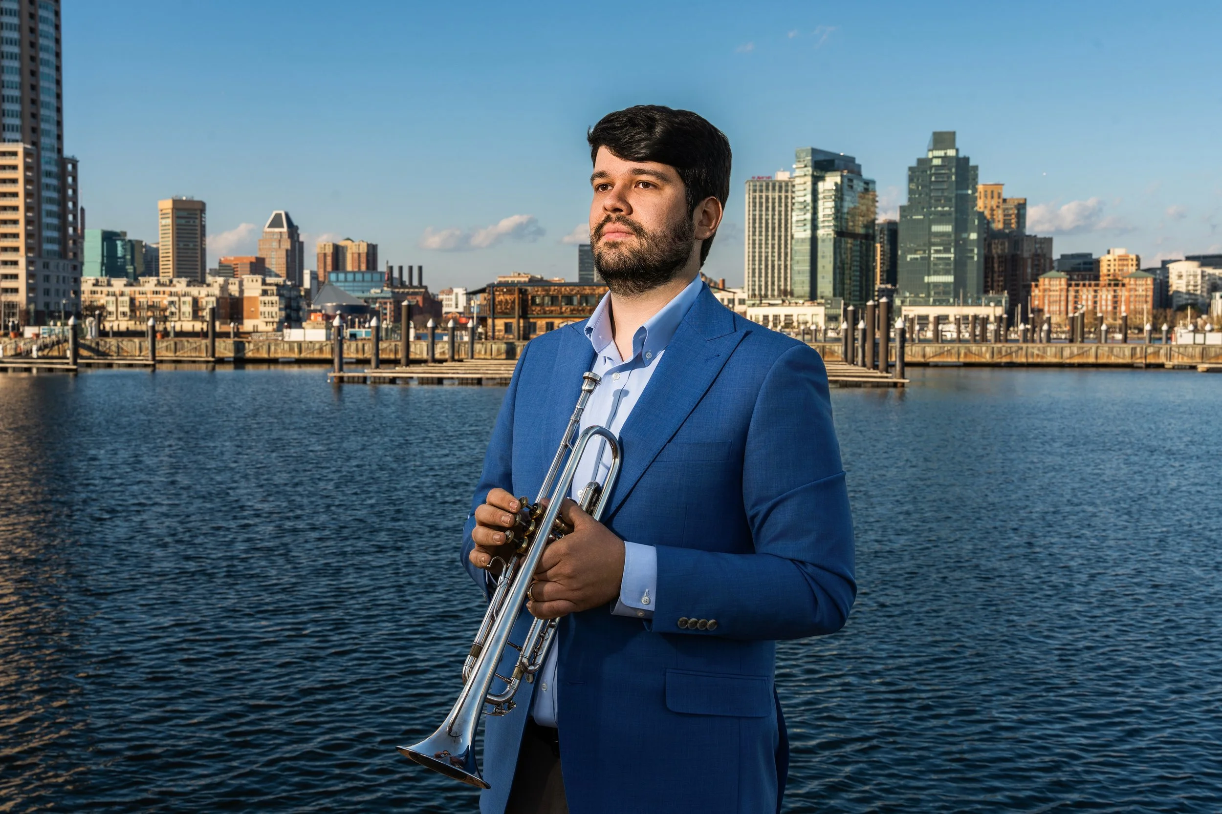 A man in a blue suit holding a trumpet stands near a body of water with a city skyline in the background.