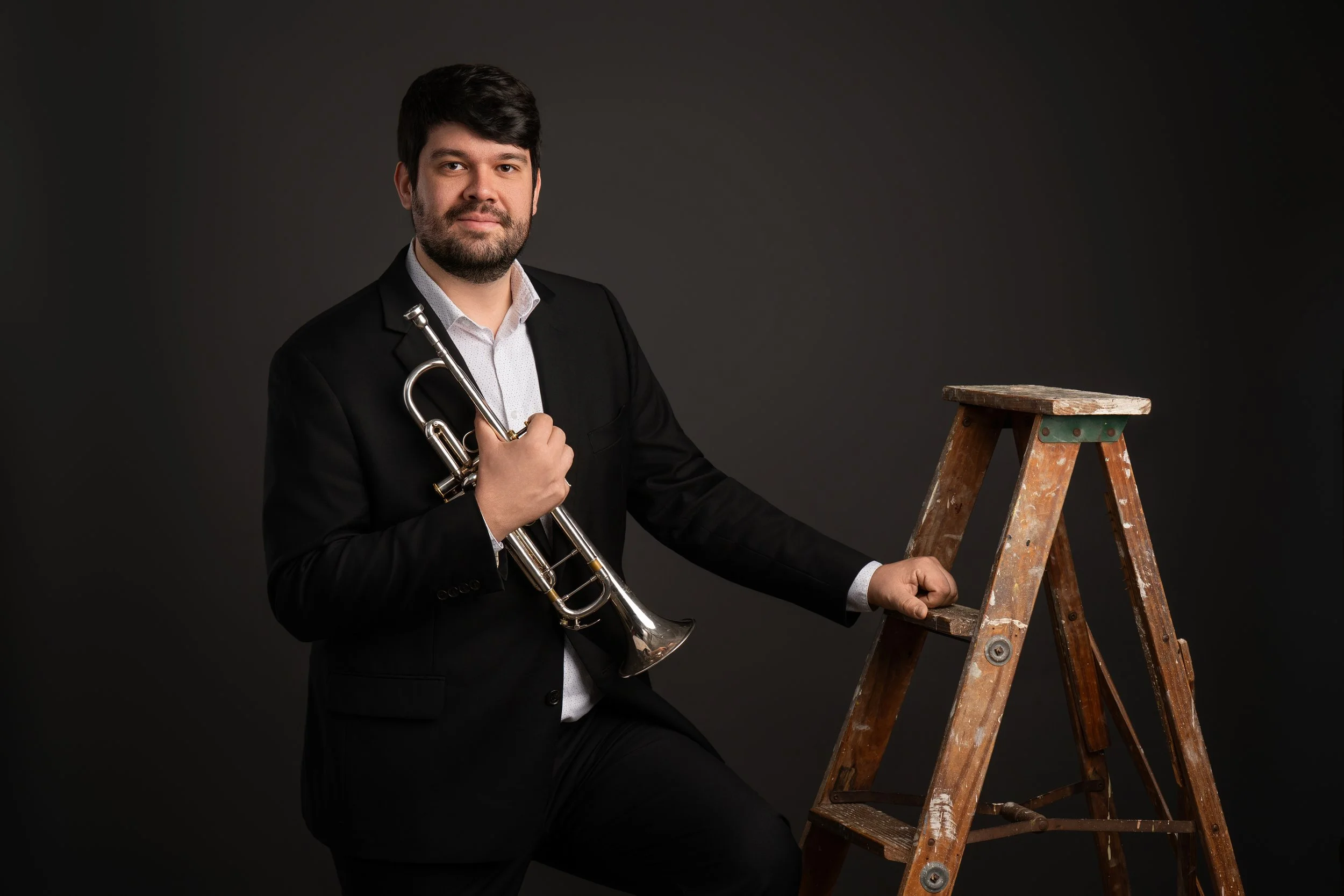 A man in a black suit holds a trumpet and rests his hand on a wooden ladder against a dark background.