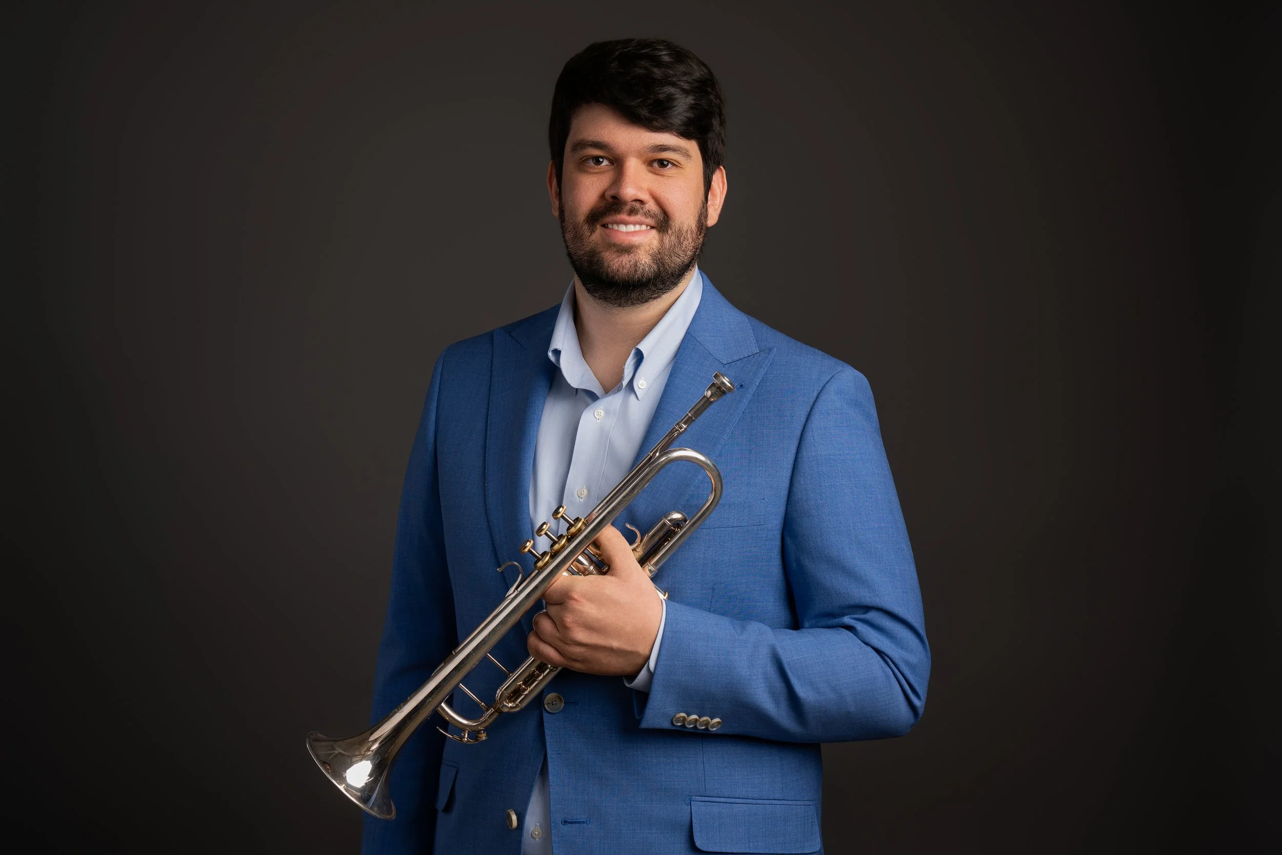 A man with dark hair and a beard wearing a blue suit jacket and white shirt, holding a trumpet, standing against a dark background, smiling.