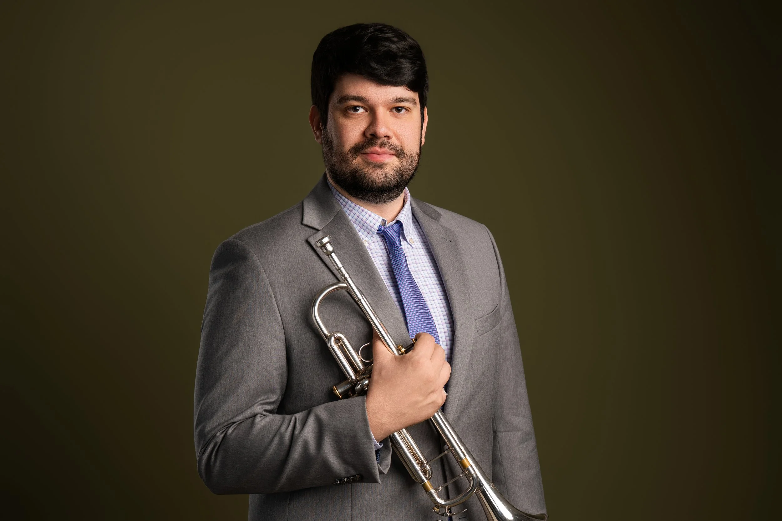 A man in a gray suit holding a silver trumpet posing against a dark olive green background.