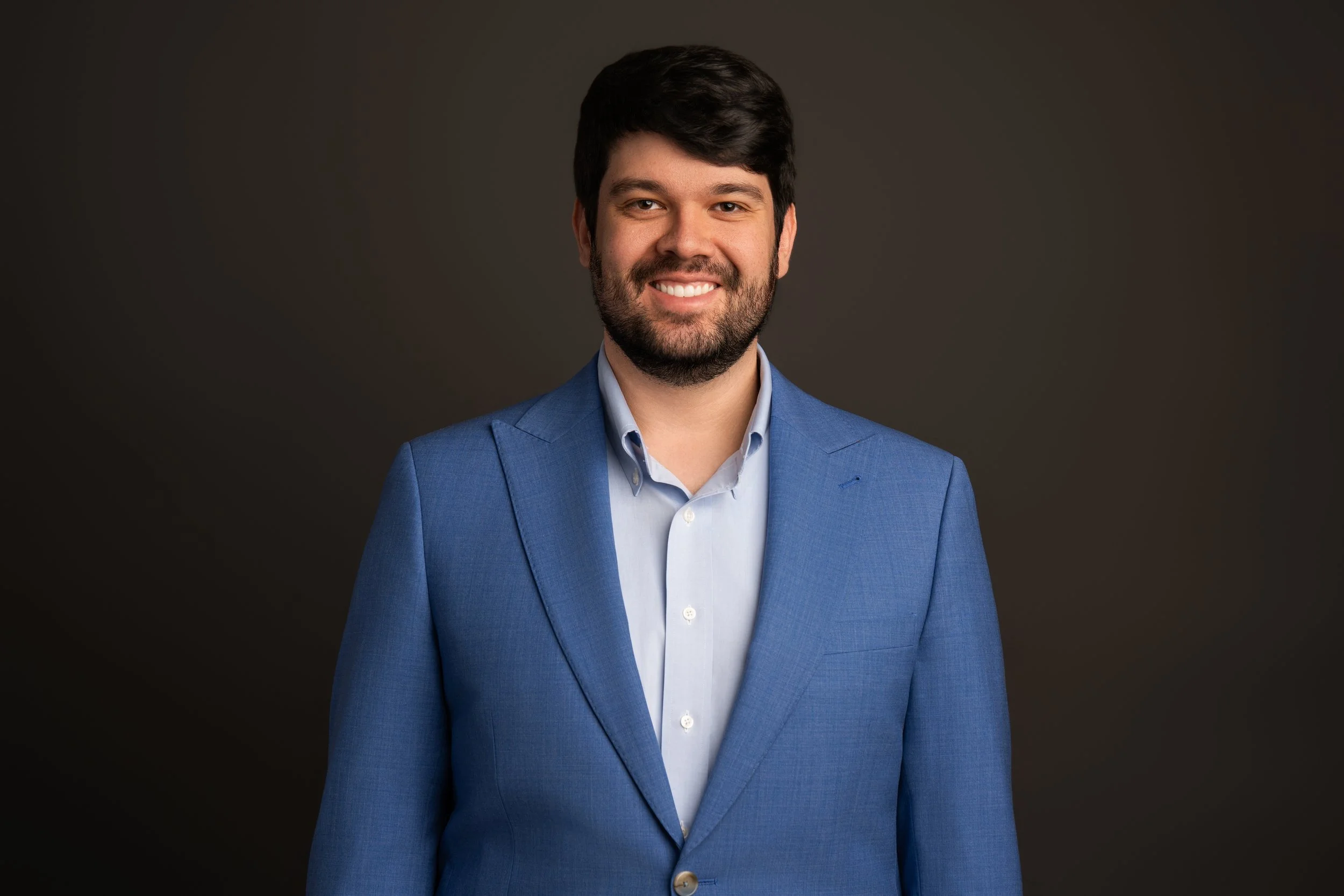 A man with dark hair and a beard wearing a blue suit and light blue shirt, smiling at the camera against a dark background.