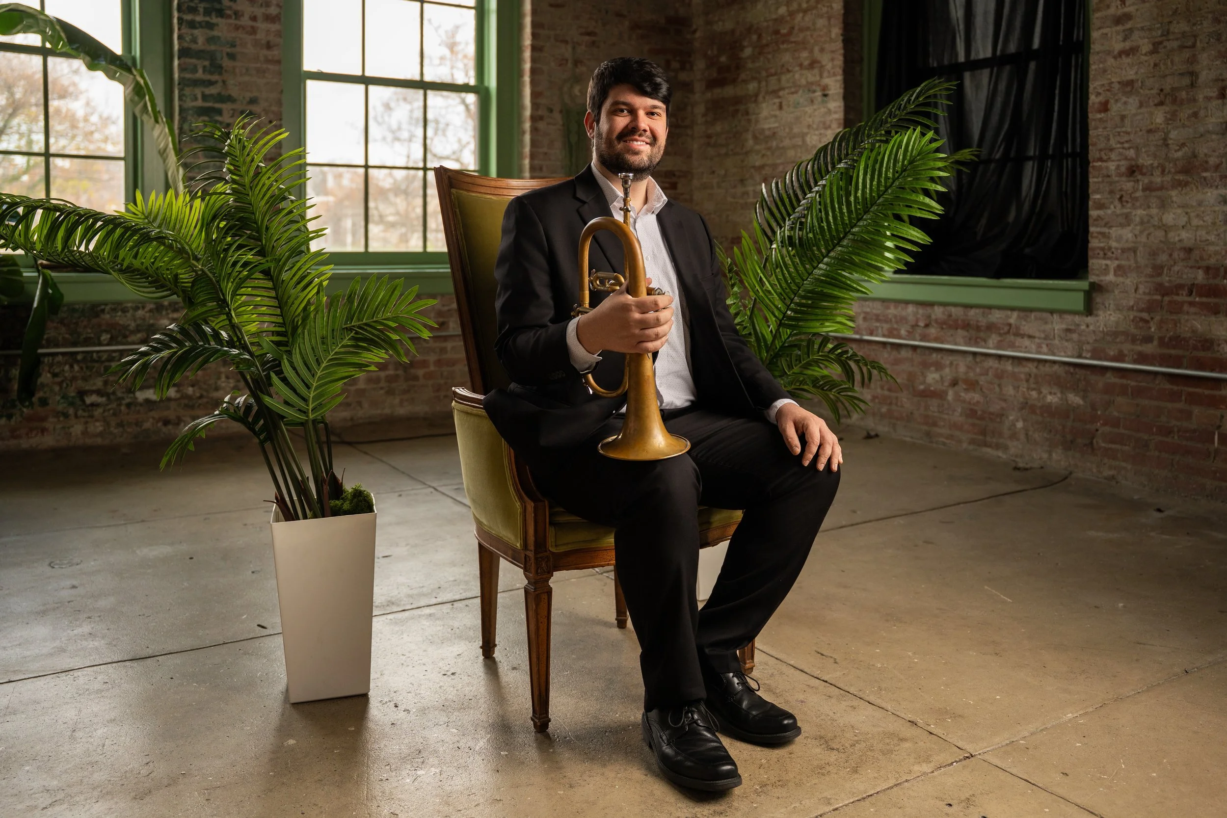A man in a black suit sitting on a vintage chair, holding a brass trumpet, smiling at the camera, with large green leafy plants on either side, in an industrial-style room with brick walls and windows.