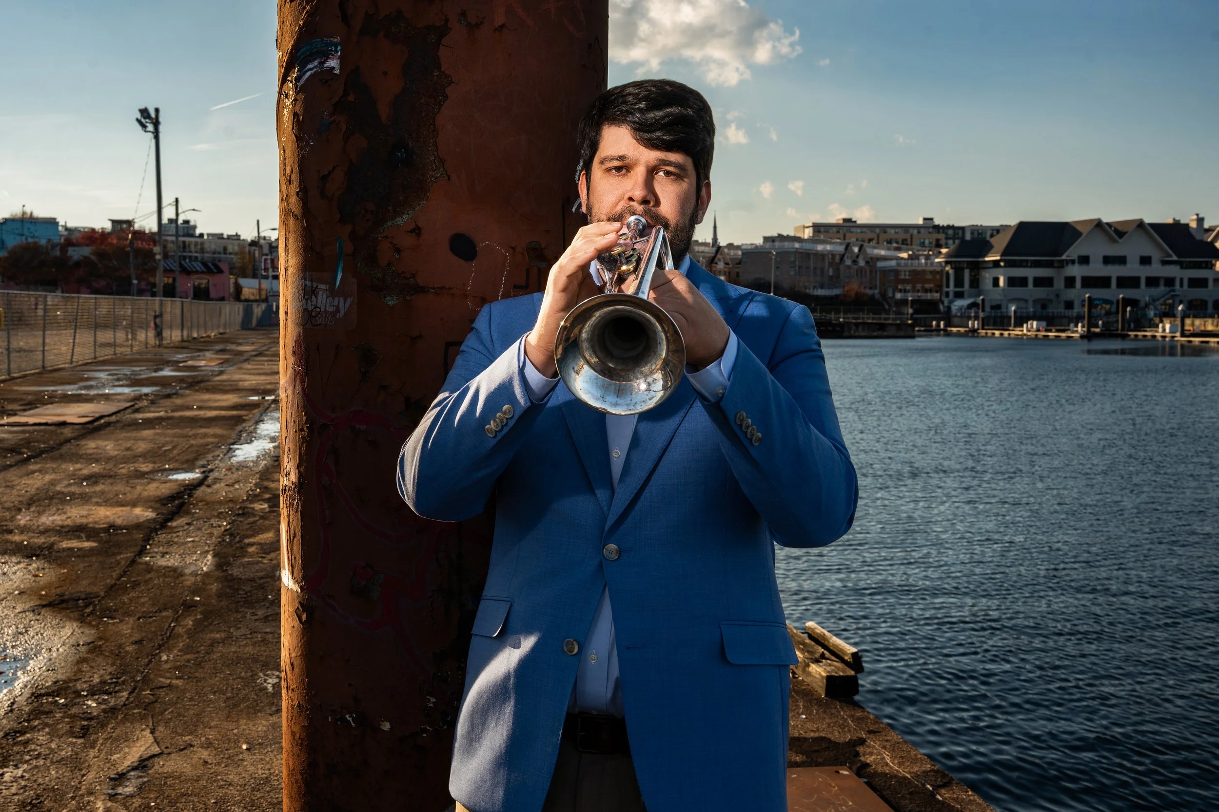 A man in a blue suit playing a trumpet by the water on a dock with buildings in the background.