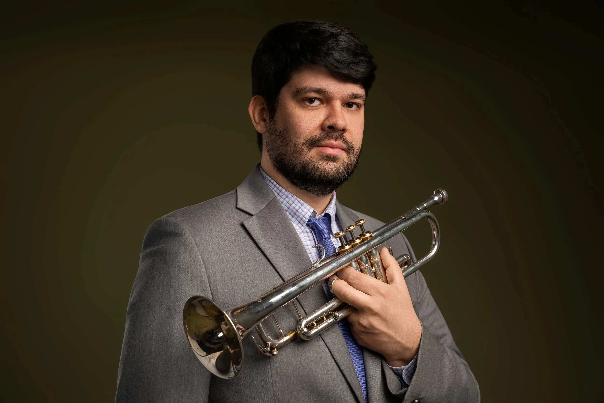 A man in a grey suit holding a trumpet against a dark background.