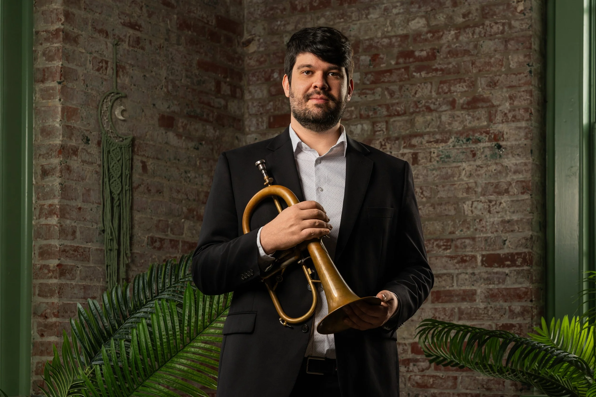 A man in a black suit holding a brass trumpet stands in a room with an exposed brick wall, green window frames, and large green plants.