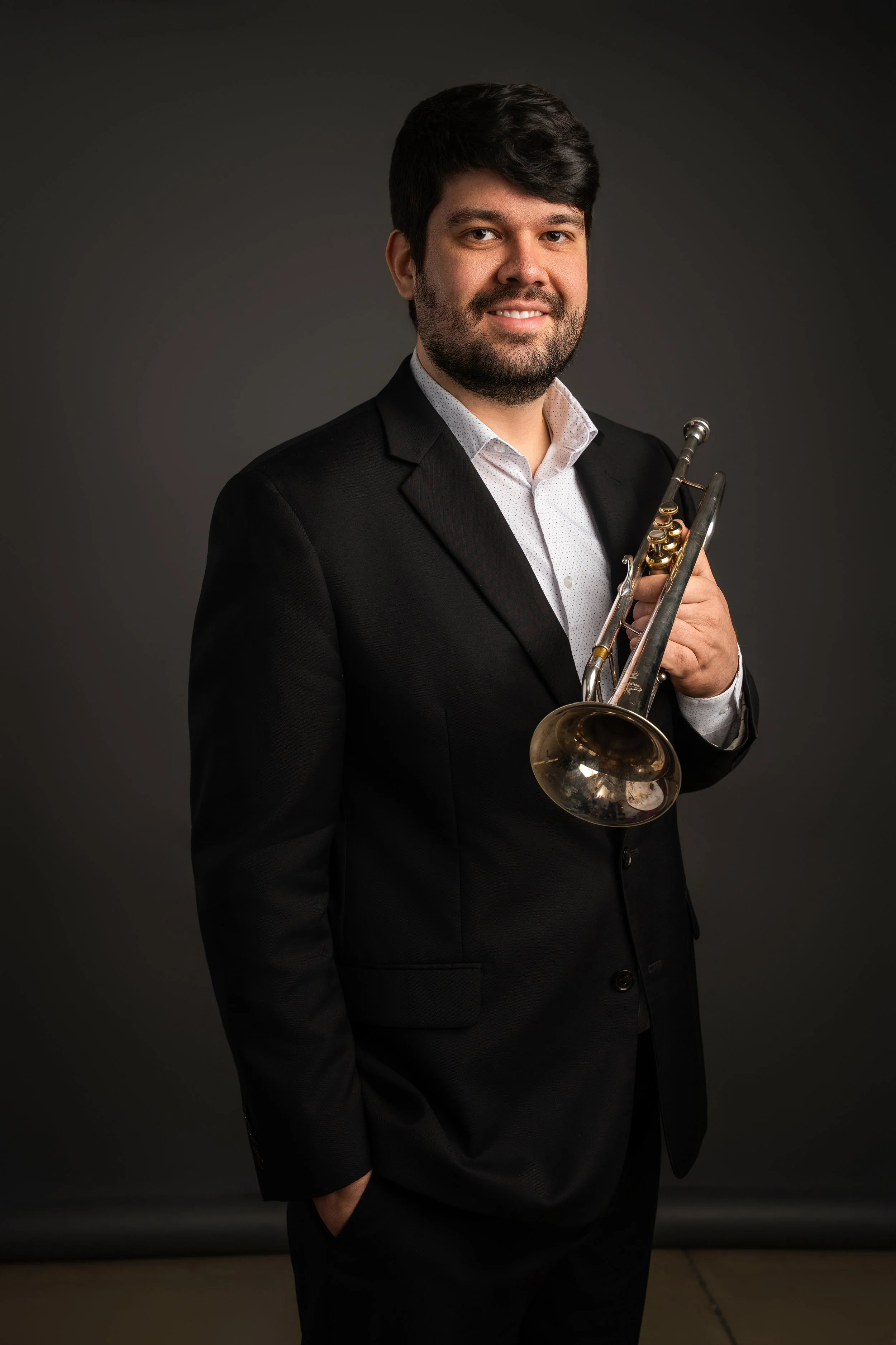 A man wearing a black suit with a white shirt, holding a trumpet, standing against a dark gray background, smiling at the camera.