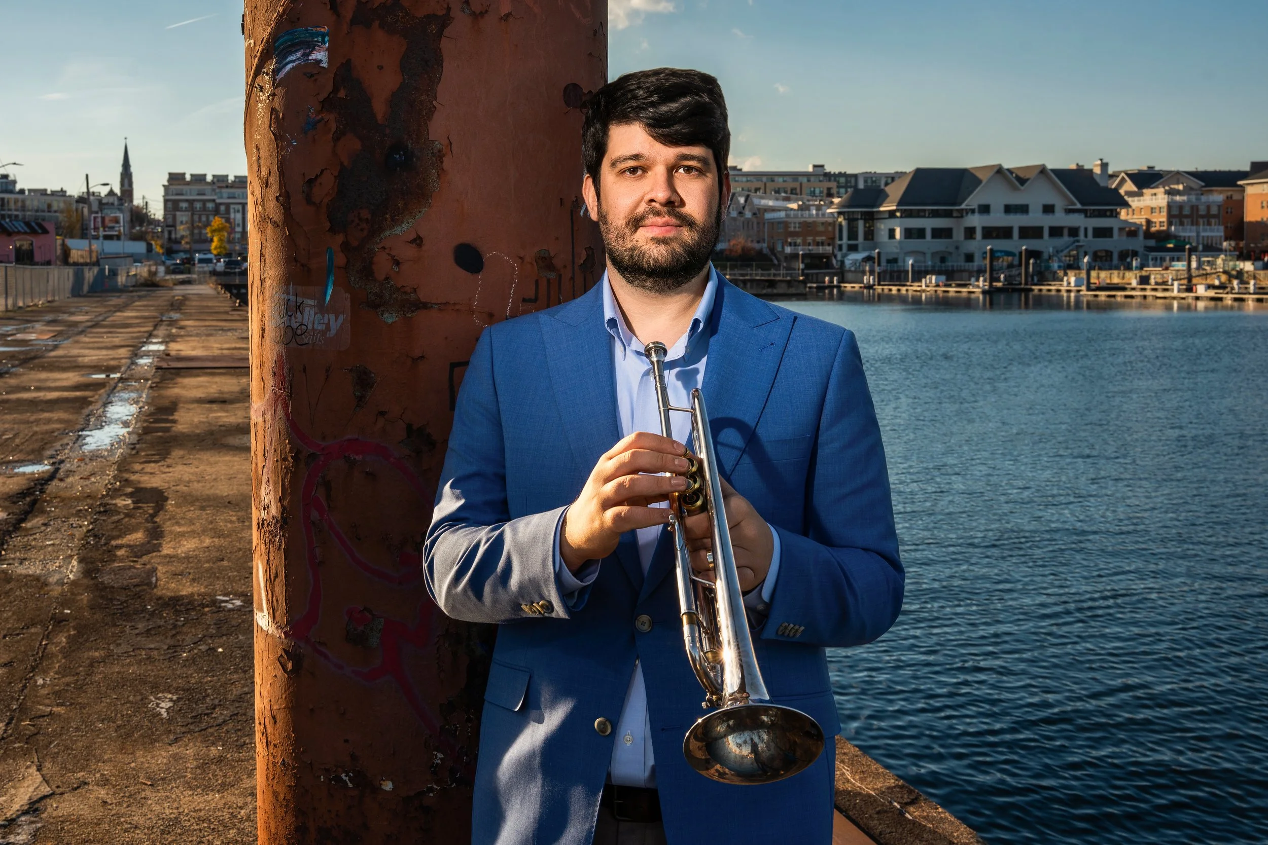 Man in blue suit holding trumpet by a waterfront with urban buildings in the background, during daytime.