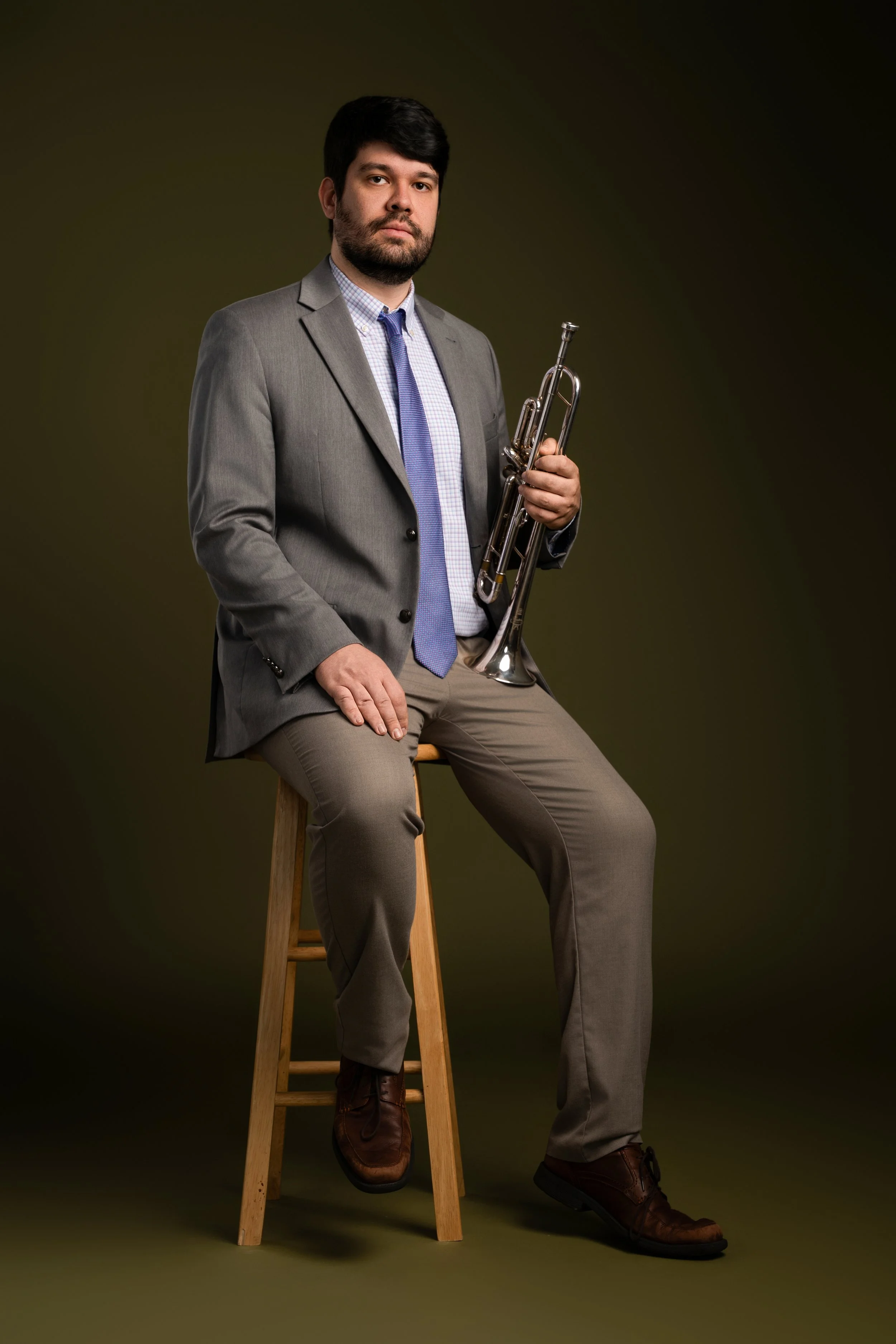 A man in a gray suit and blue tie sitting on a wooden stool, holding a trumpet, against a dark background.