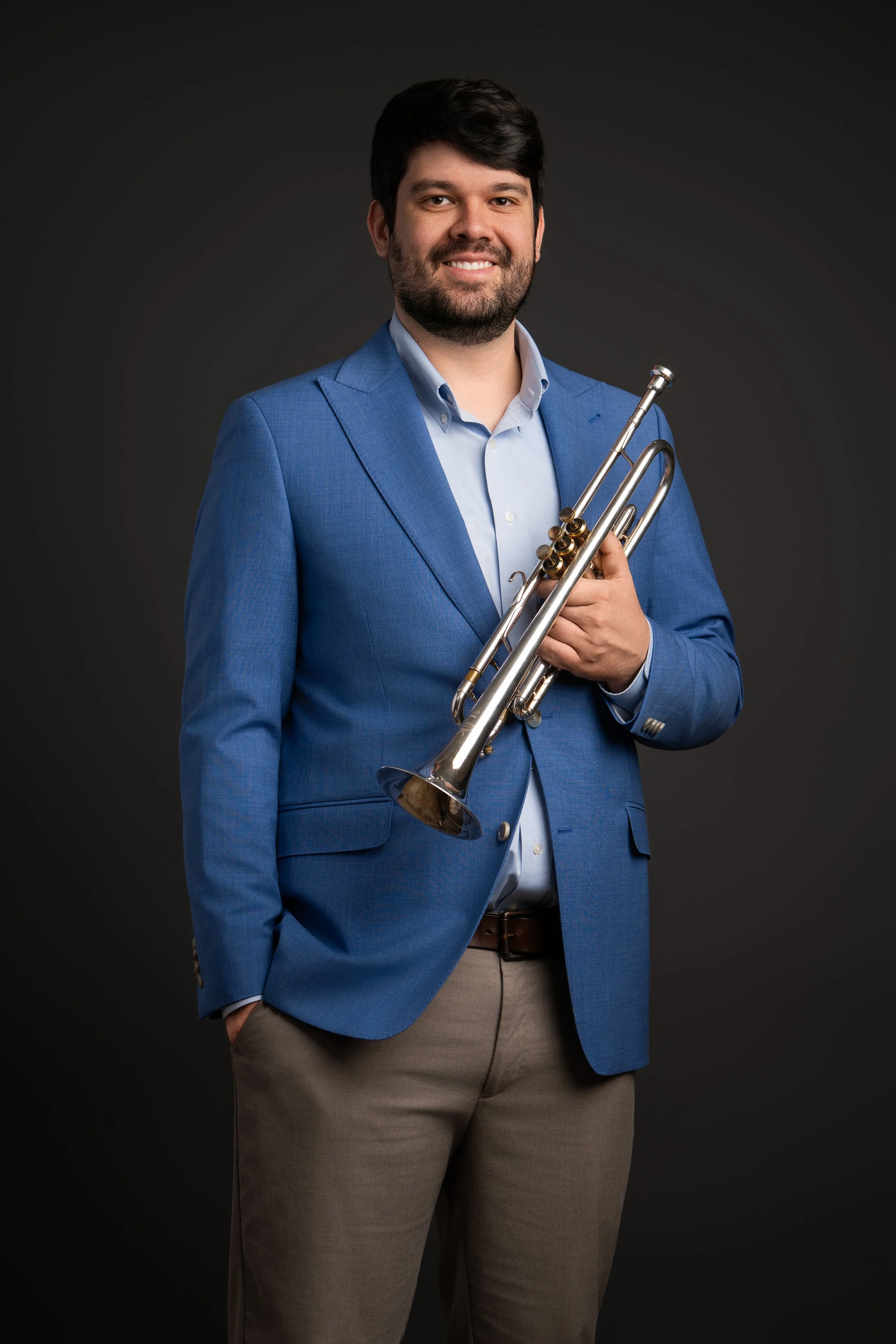 A man with dark hair and a beard wearing a blue blazer, light blue shirt, and khaki pants, holding a trumpet, standing against a dark background.
