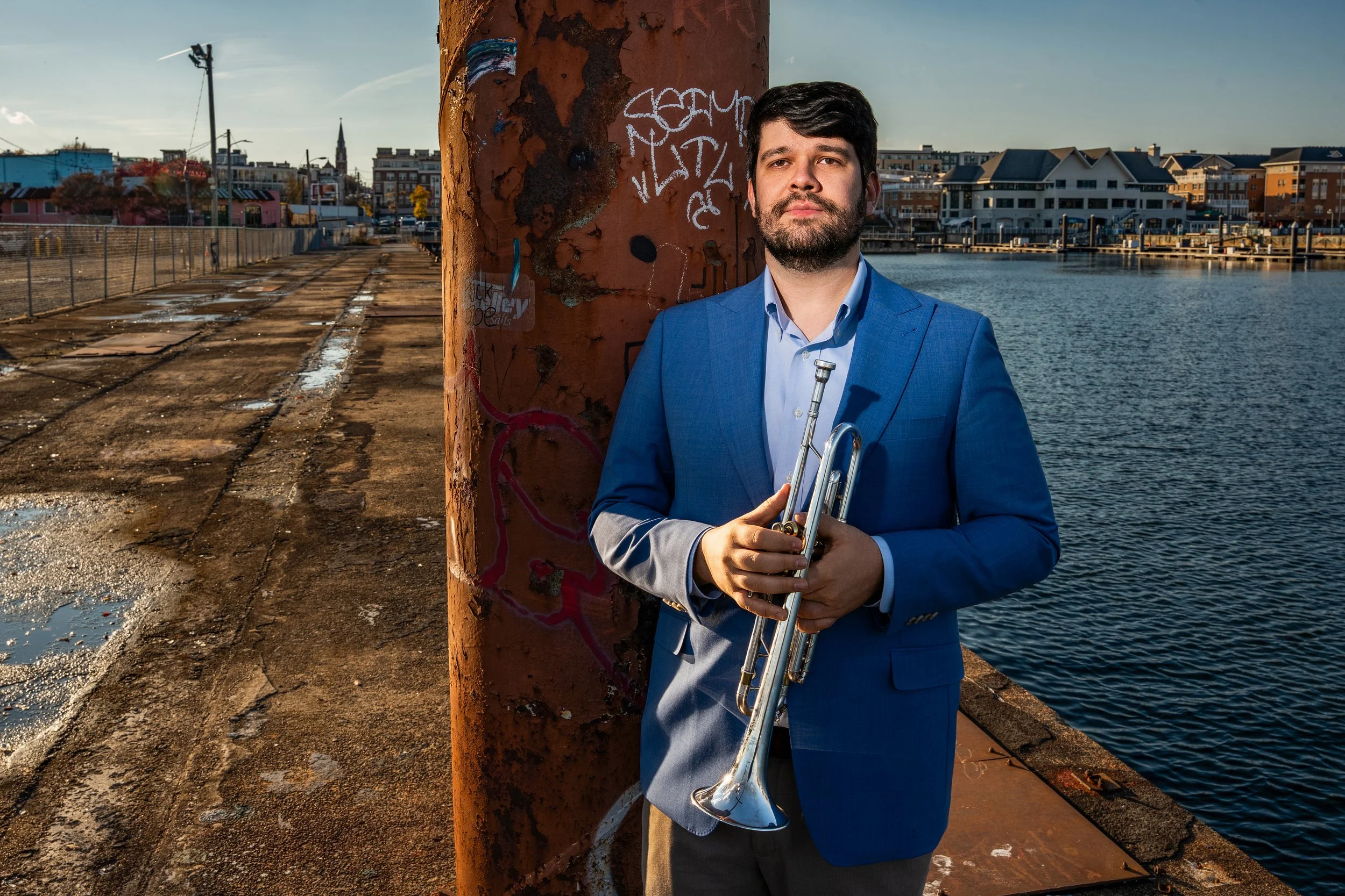 A man in a blue suit holding a trumpet while standing next to a rusty pole by the water in an urban area.