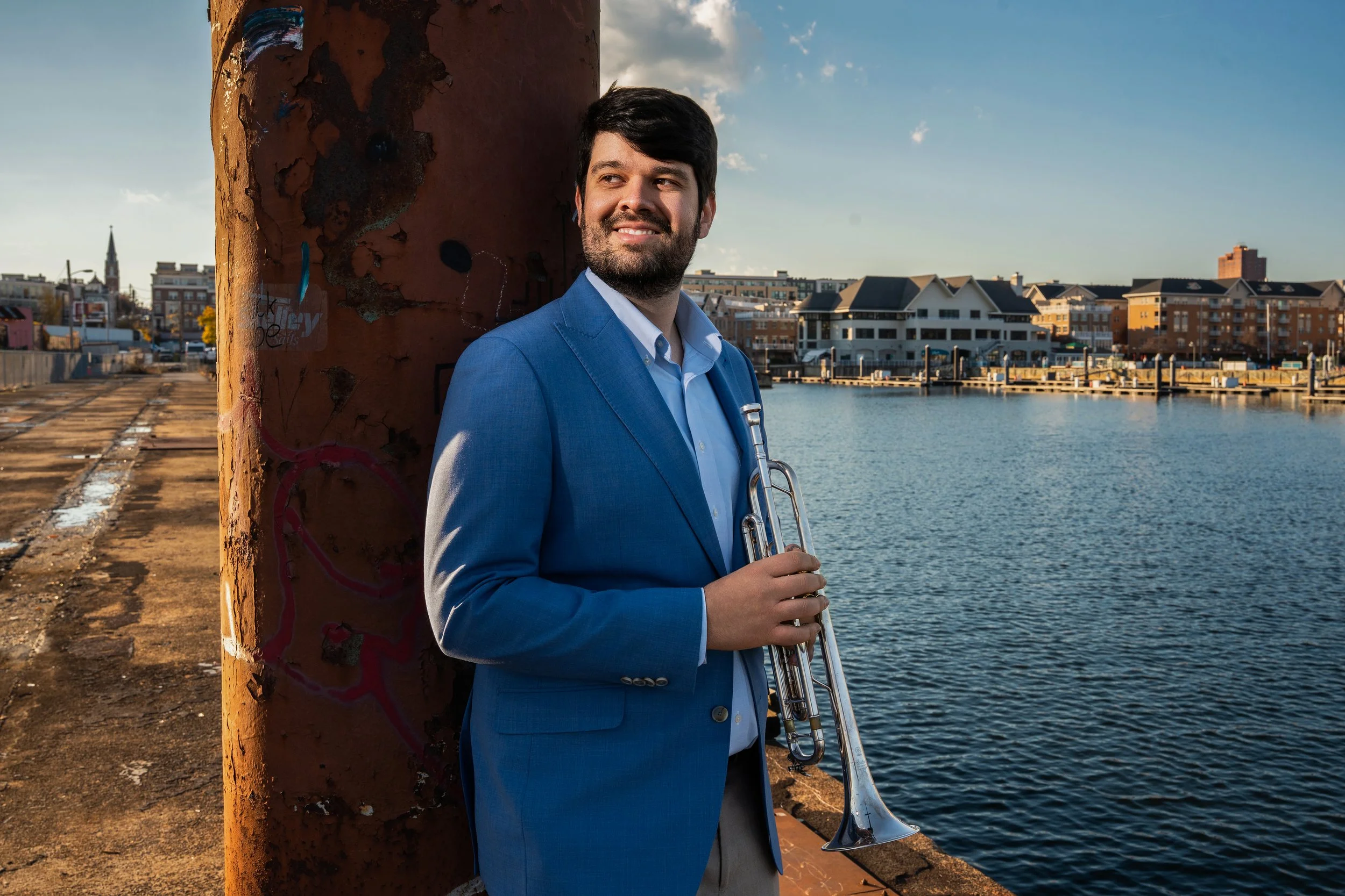 A man in a blue suit holding a trumpet, standing by a dock with water and buildings in the background during sunset.