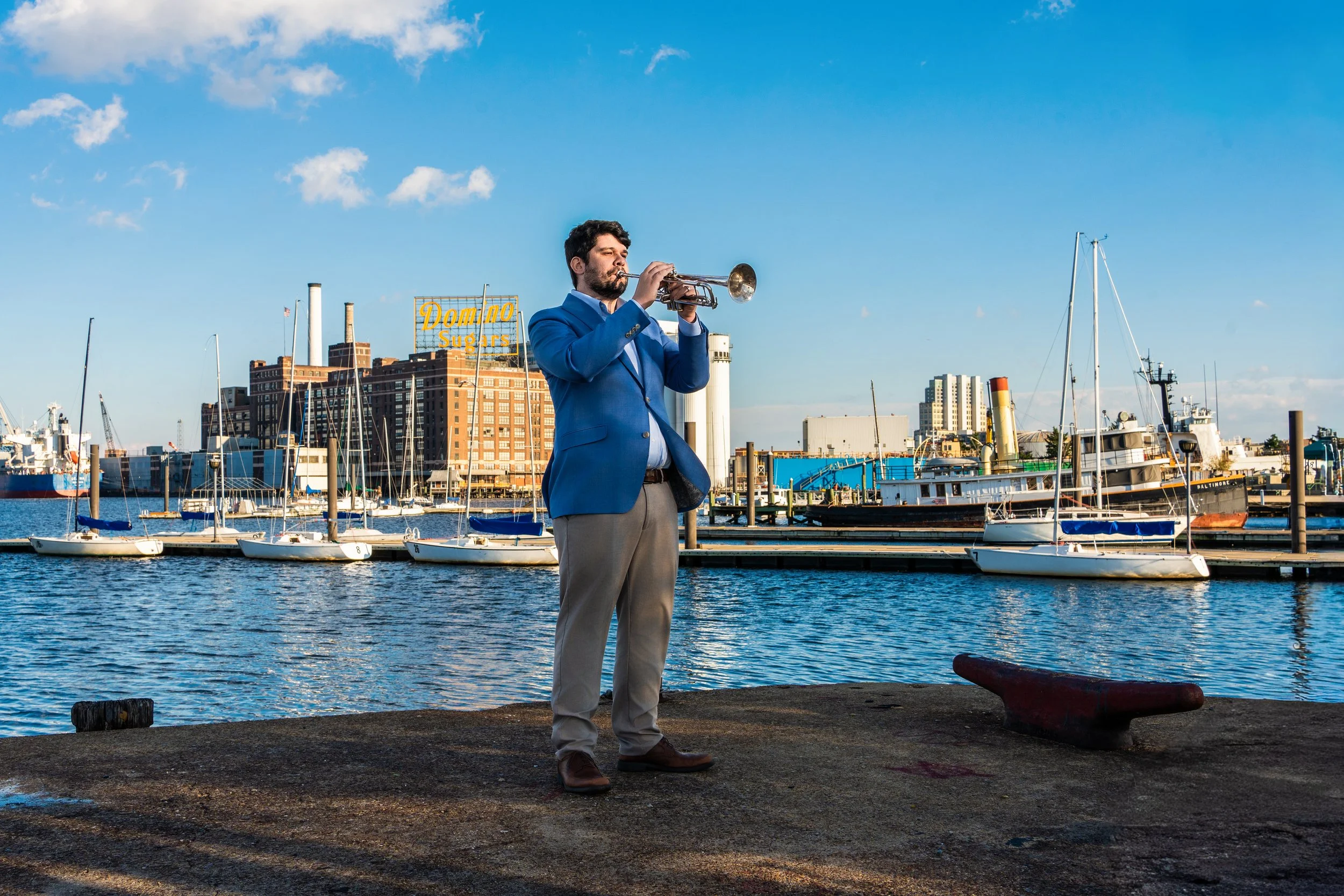 A man in a blue blazer playing the trumpet by a harbor with sailboats and an industrial city skyline in the background.