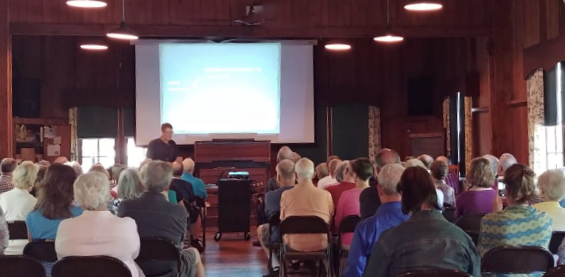 A person giving a presentation to a seated audience in a wood-paneled room with a large screen displaying a slide.