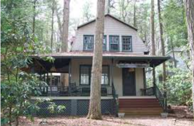 A two-story house surrounded by trees with a large front porch, stairs leading up to the entrance, and a black railing.