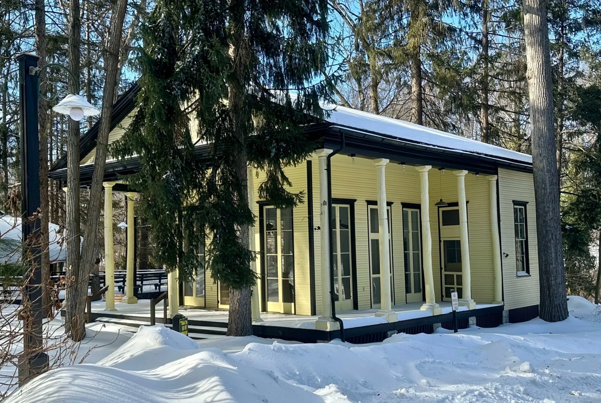 A yellow house with tall windows and a porch, surrounded by snow and trees in winter.