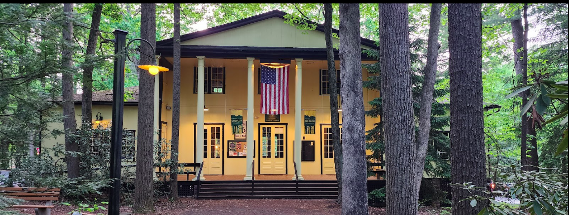 A yellow building with white columns and a porch, surrounded by tall trees in a forest setting. An American flag hangs on the front of the building.