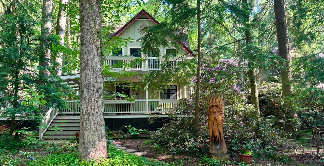 A two-story house with a front porch, surrounded by greenery and trees, with a wooden sculpture of a face in the garden.