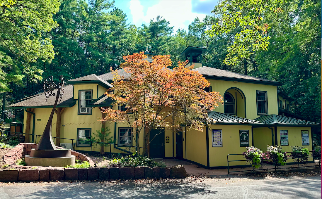 Yellow building with black trim and metal roof, surrounded by trees and bushes, with a sculpture in the foreground.