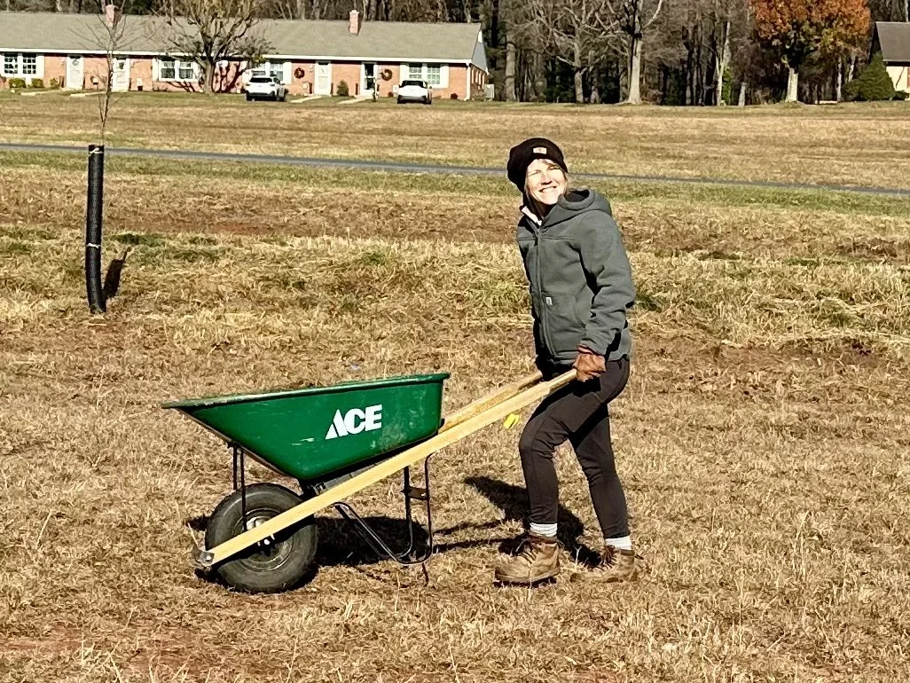 A woman in outdoor clothing and boots pushing a green wheelbarrow in a grassy field.