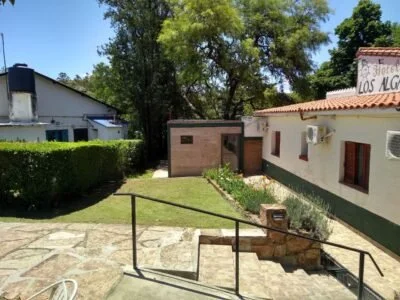 Entrada a un monumento o sitio con caminos de piedra y barandales, rodeado de árboles y edificios, en un día soleado.