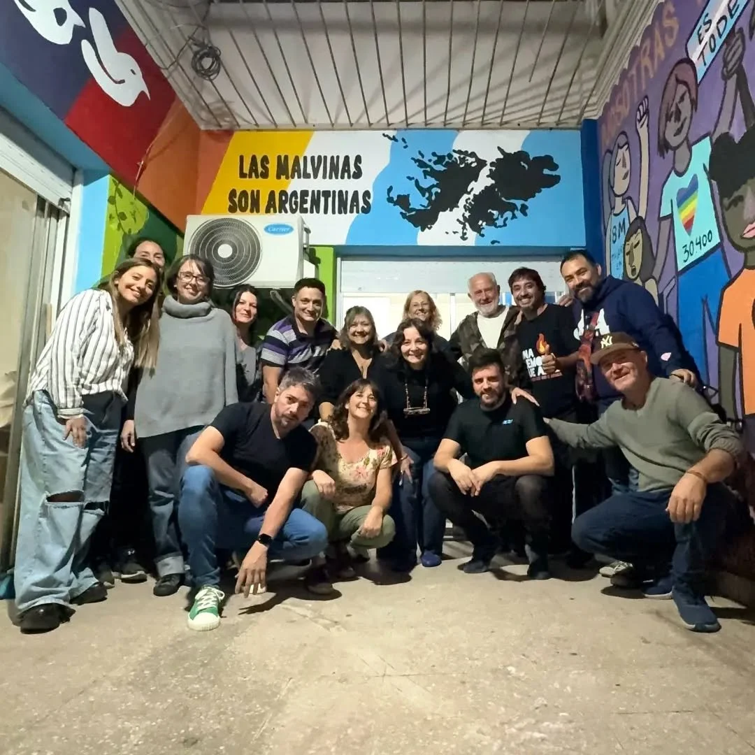 Un grupo de personas sonrientes posando en una sala con paredes pintadas con mapas y textos en español, incluyendo uno que dice 'Las Malvinas son argentinas'.