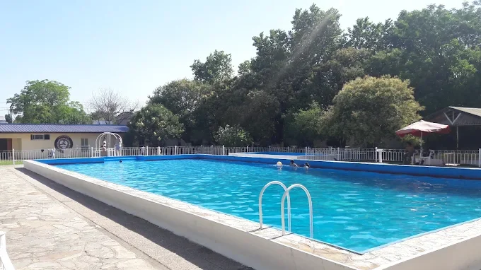 Piscina al aire libre con agua azul, rodeada de una cerca blanca, en un área con árboles y sombra, en un día soleado.