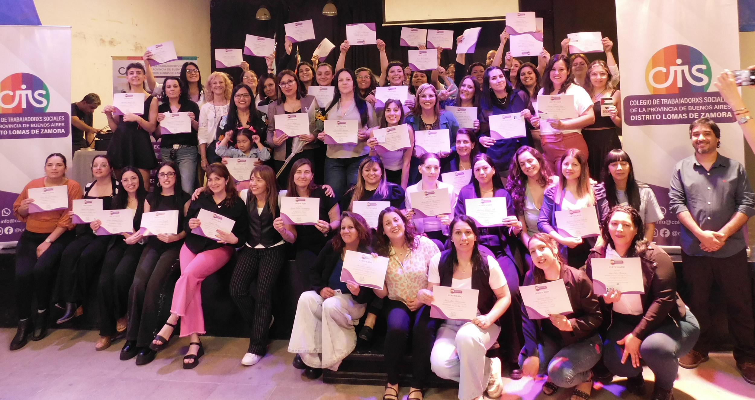 Grupo de personas en ceremonia de entrega de certificados, algunos posando con sus reconocimientos y otras levantando diplomas, en un evento organizado por el Colegio de Trabajadores Sociales de la provincia de Buenos Aires.
