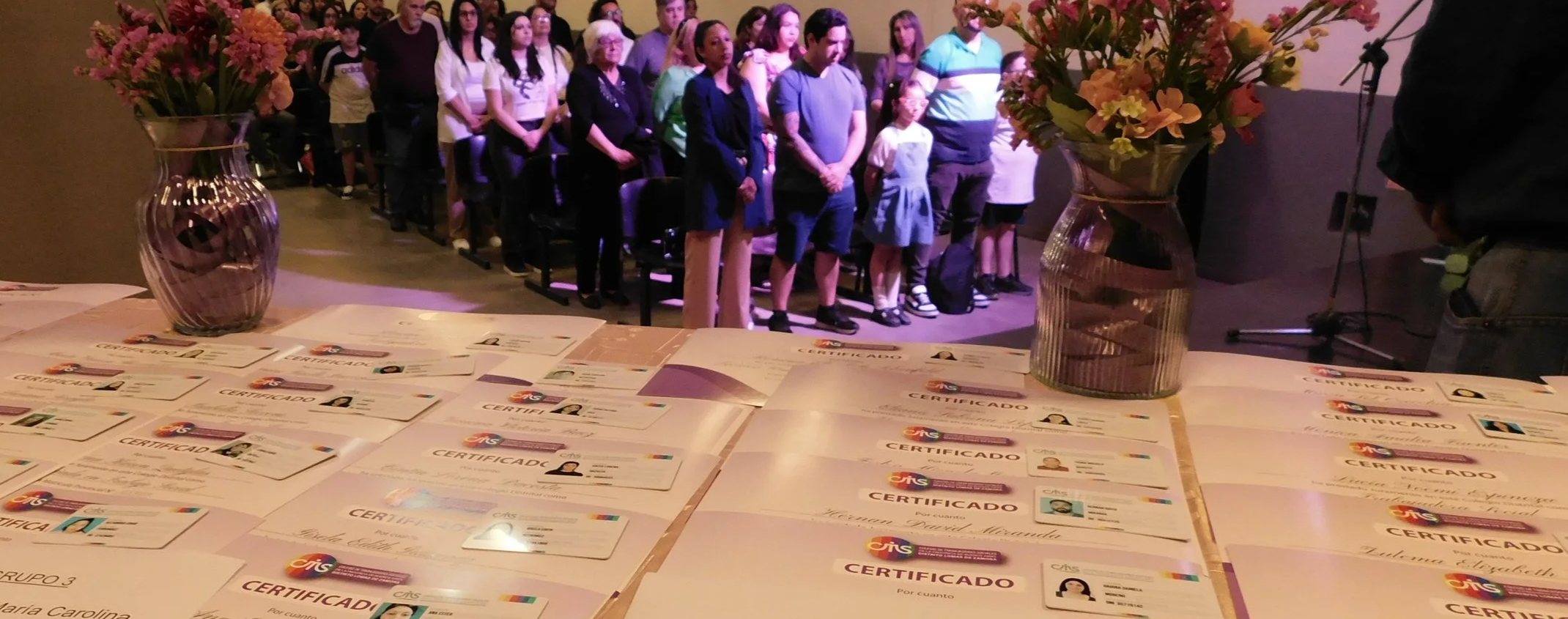 Personas de diferentes edades en una ceremonia, con certificados sobre una mesa en primer plano y un grupo de personas de pie en el fondo, algunos con las manos juntas, en un espacio interior con iluminación cálida.