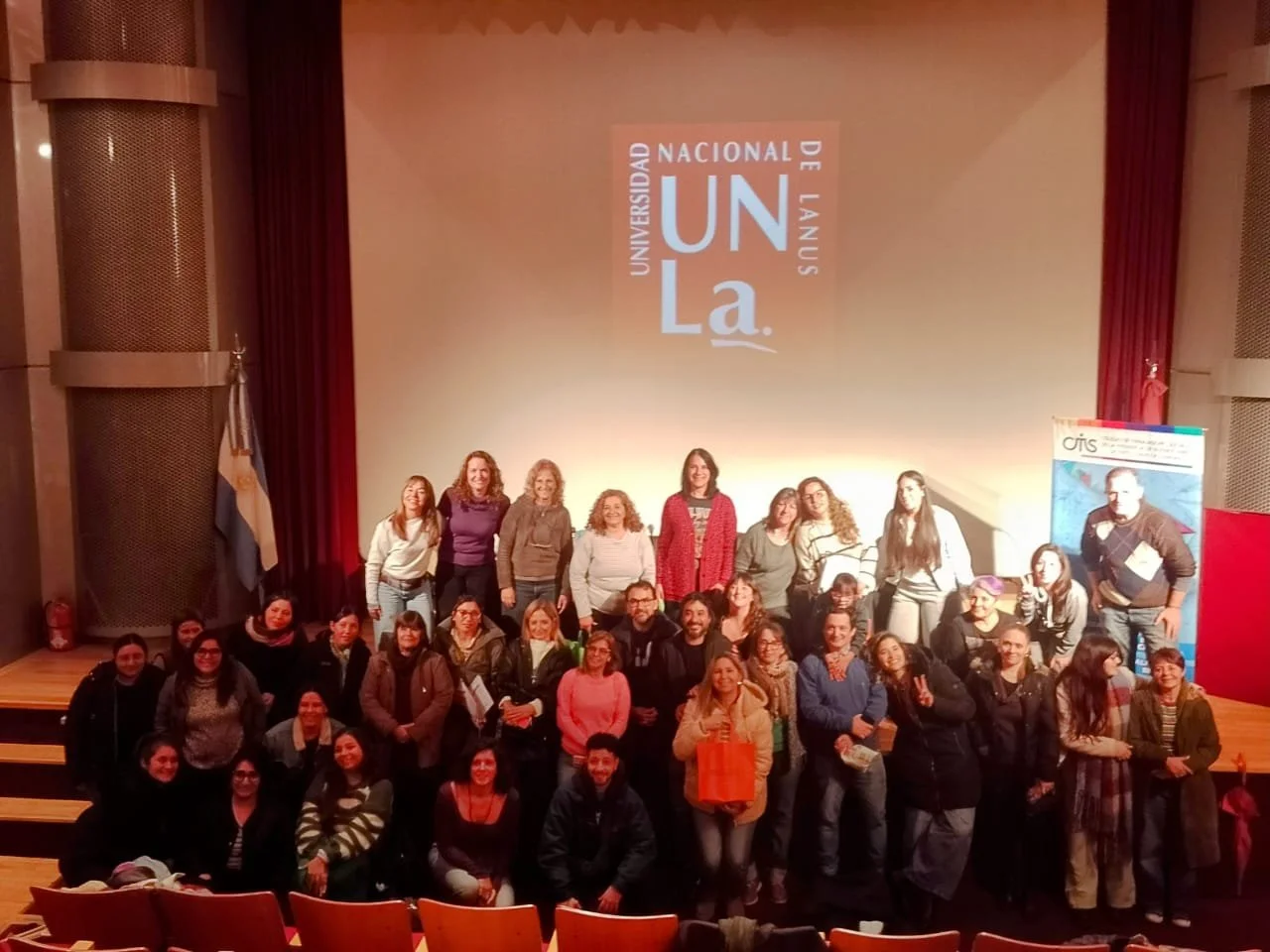 Grupo de personas en un escenario de teatro frente a una pantalla con el logotipo de la Universidad Nacional de Laus en Argentina, algunos posando y otros conversando.