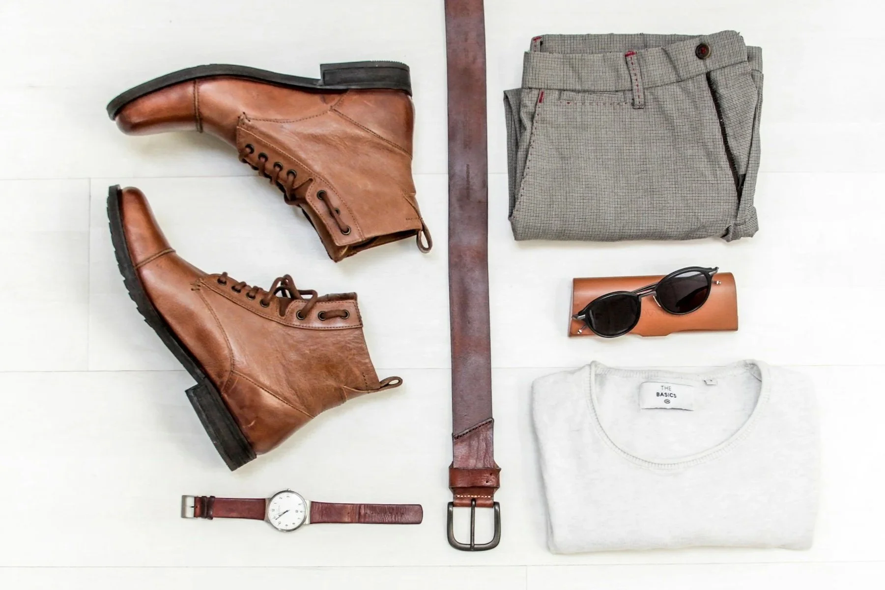 Zapatos de cuero marrón, pantalones de vestir grises, gafas de sol negras, cinturón de cuero marrón, reloj de pulsera con correa de cuero, camiseta gris con etiqueta "The Basics"