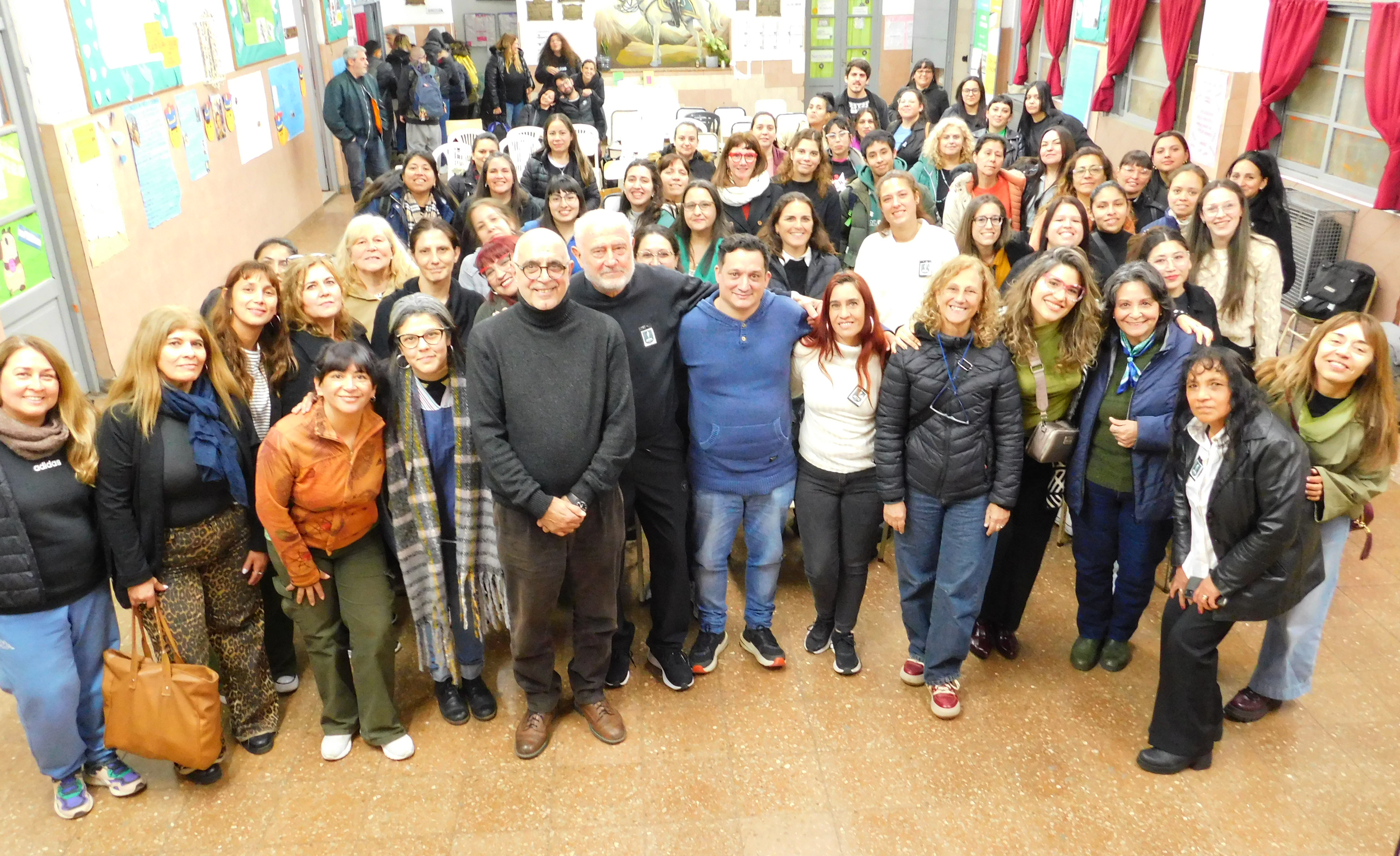 Grupo de personas posando en una sala de clases o salón comunitario, algunos sonrientes y abrazados, con una pared decorada con carteles y cuadros de fondo.
