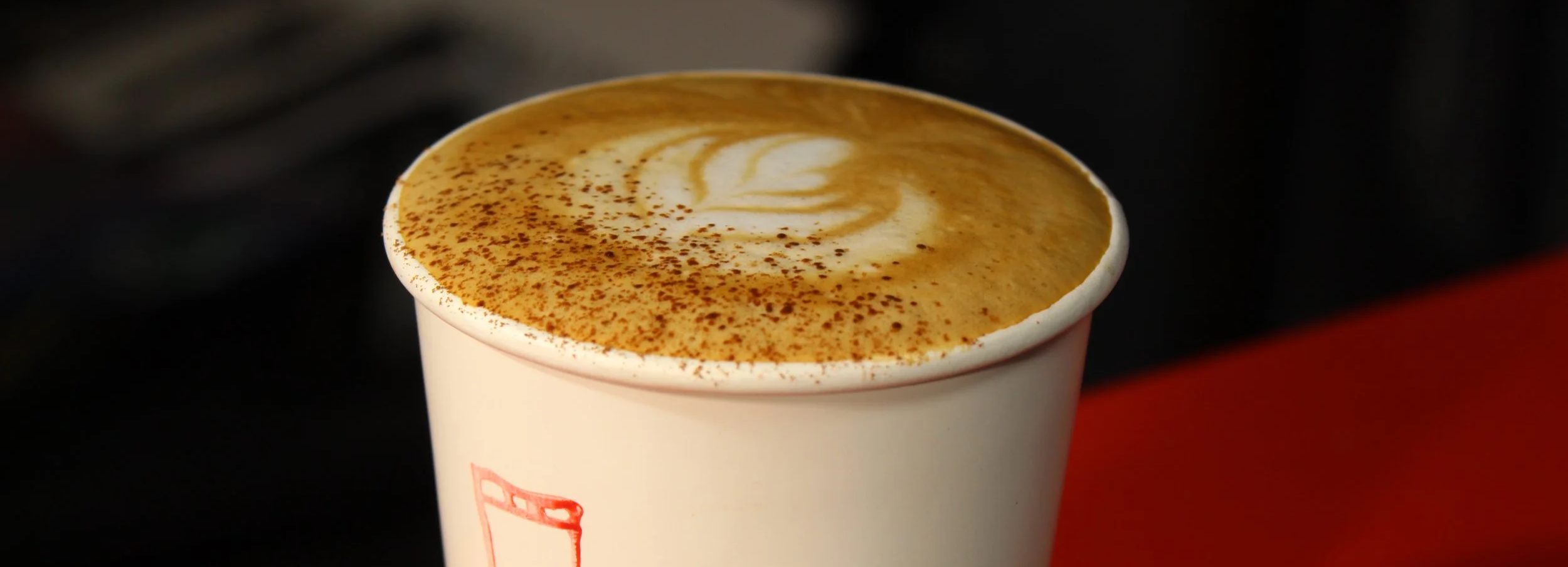 A paper cup filled with a frothy coffee beverage topped with cinnamon powder, with latte art visible on top.