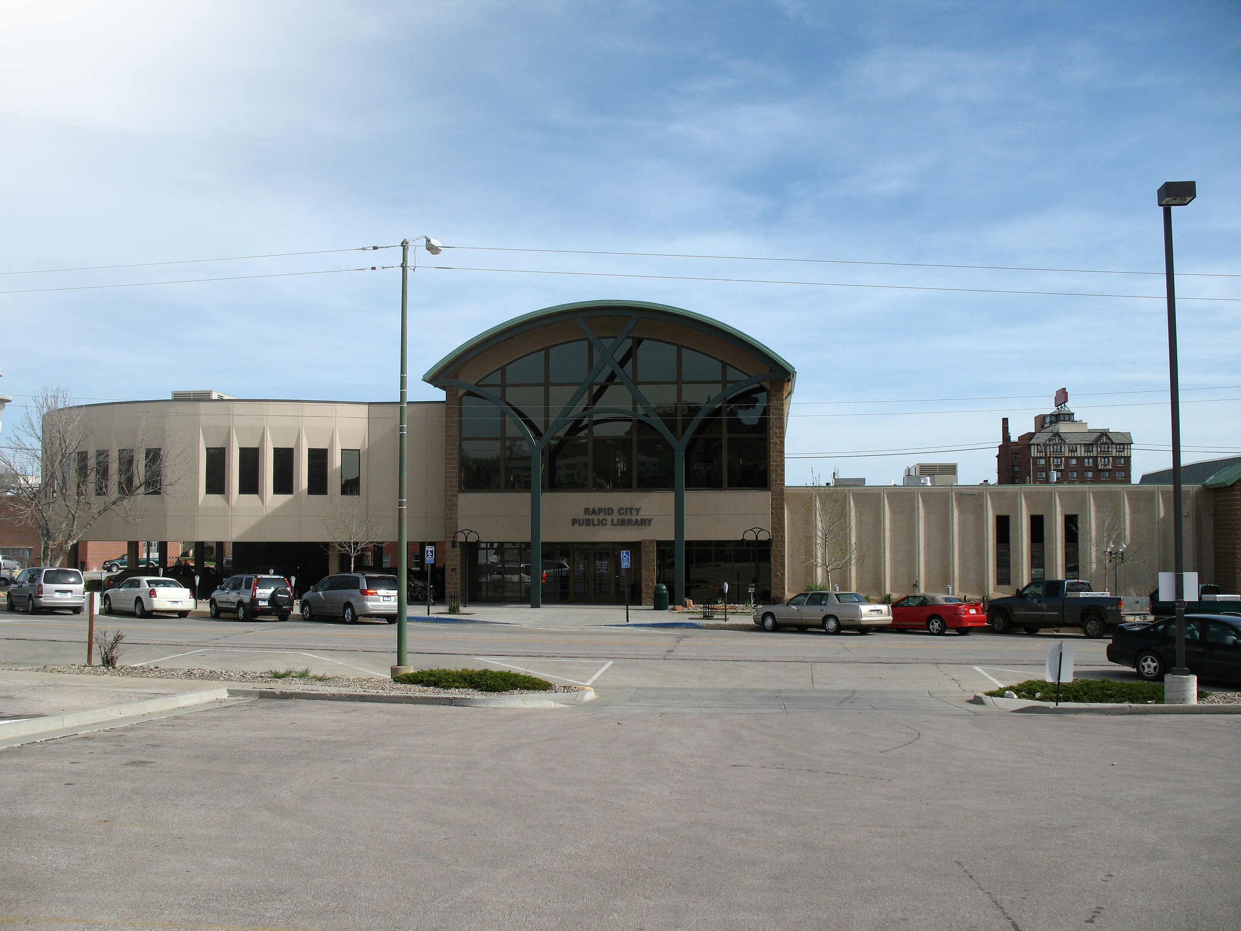 Front view of Rapid City Public Library building with parking lot in front and a few cars parked. Clear sky above with some utility wires and a streetlight.