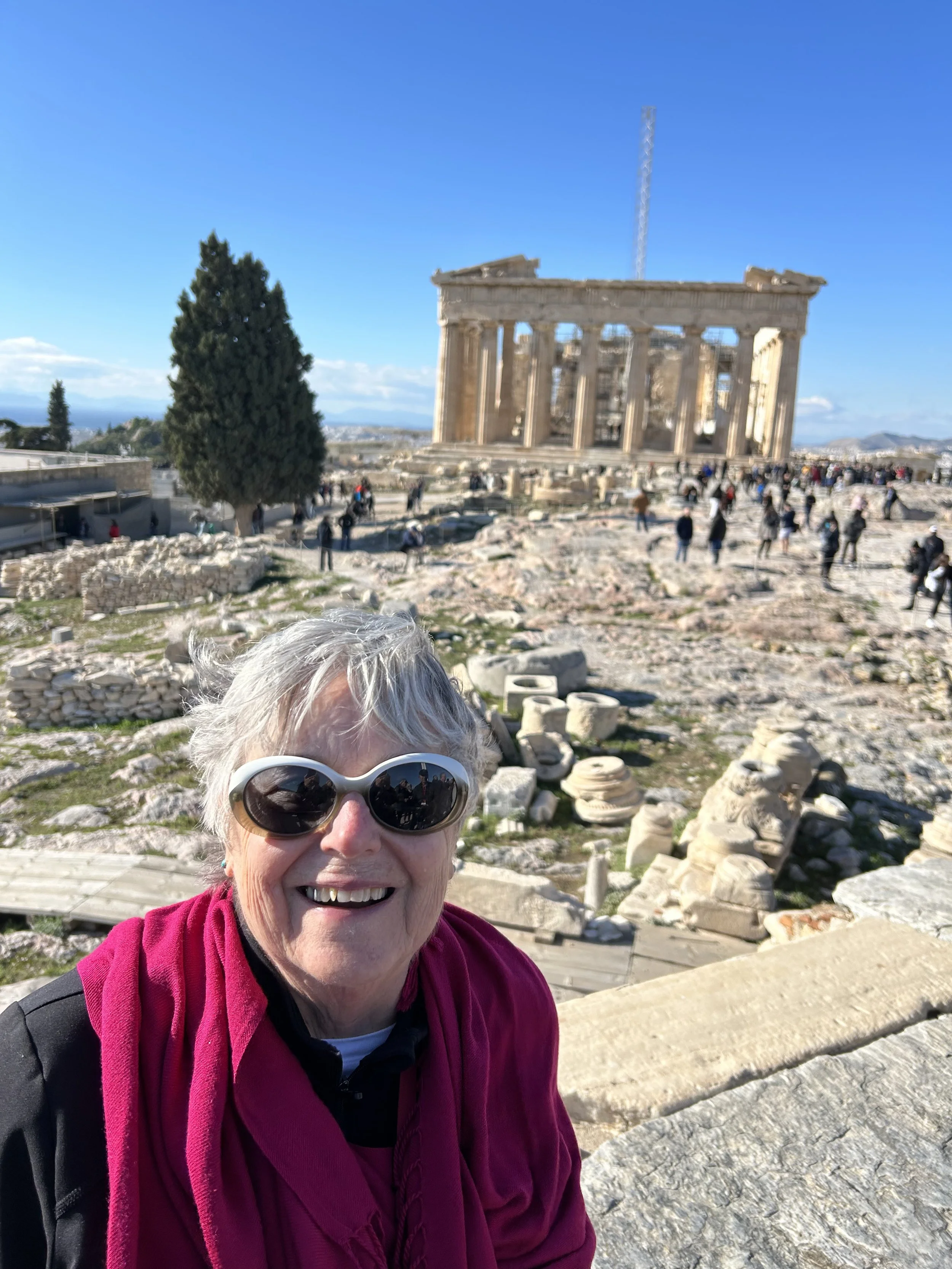 A woman wearing sunglasses and a red jacket taking a selfie at the ancient Acropolis in Athens, Greece, with the Parthenon temple in the background on a clear, sunny day.